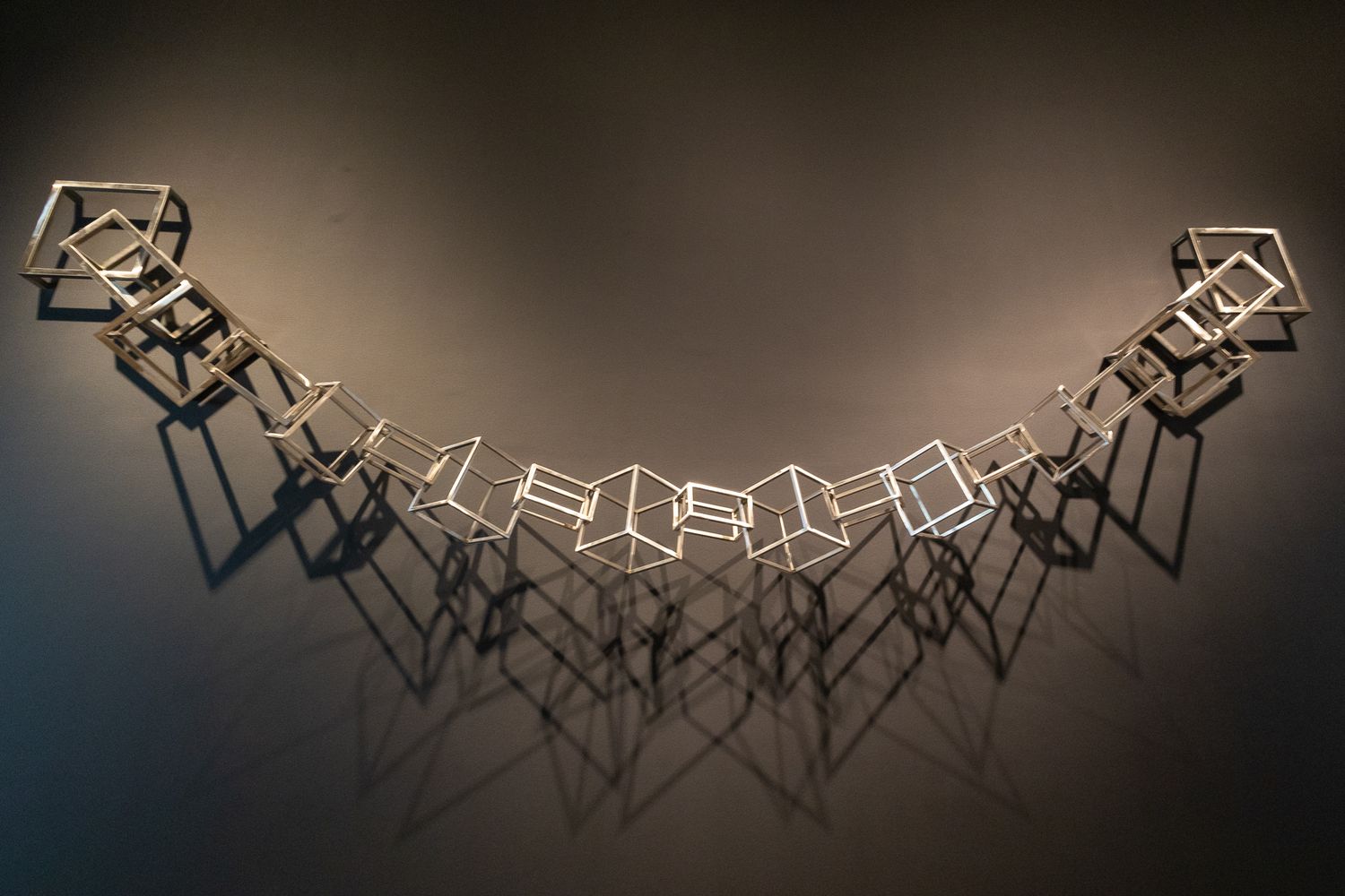 Geometric wire cube sculpture forming an arc shape casting shadows on a gray wall.