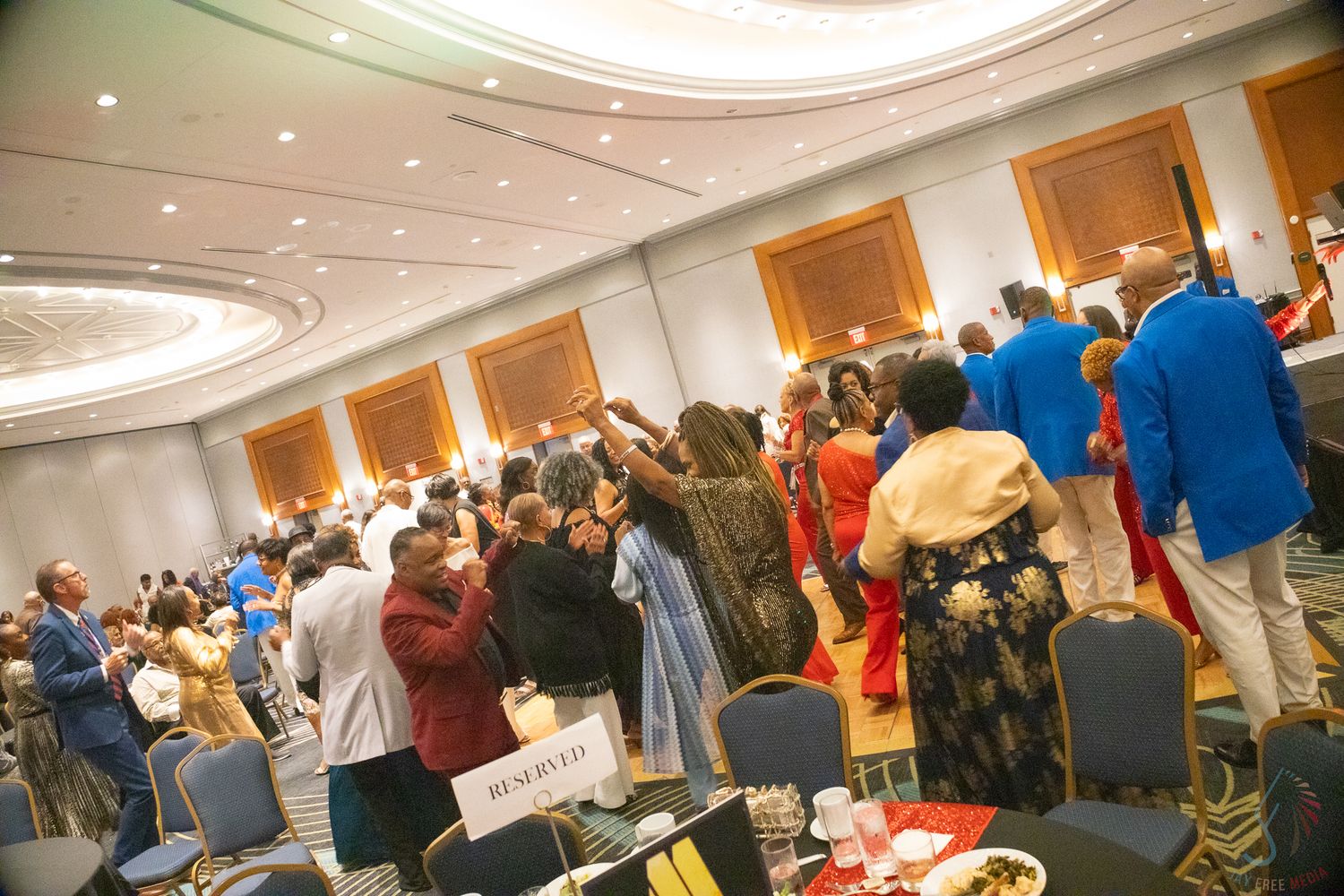 Diverse crowd of guests raising hands in celebration inside a decorated hotel ballroom.