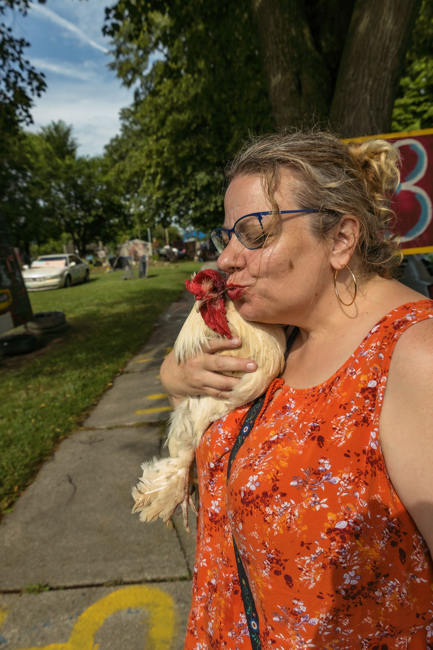 Woman in orange floral dress kissing a white rooster outdoors on a sunny day near a tree-lined sidewalk.
