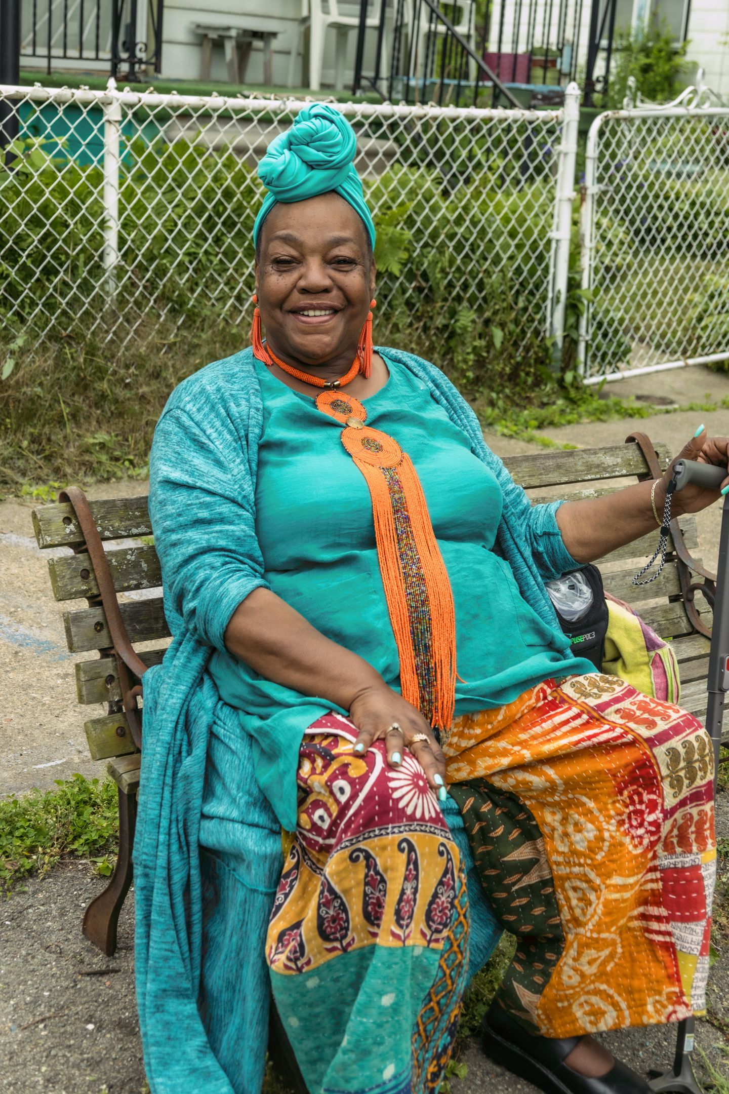 Smiling woman in teal outfit with orange accessories and colorful patterned skirt sits on bench outdoors.