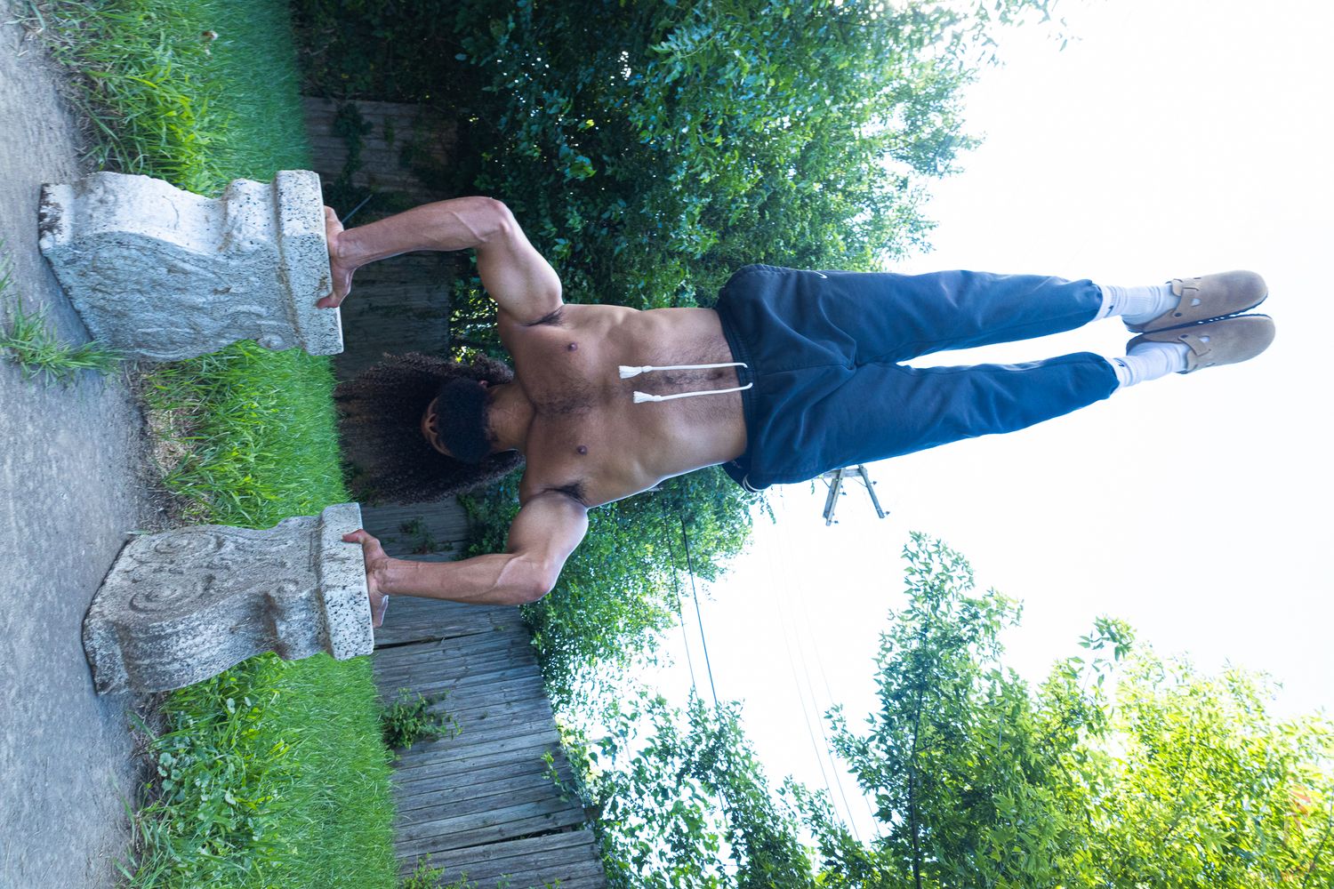 Shirtless person doing a handstand on stone sculptures in a lush green outdoor park setting.