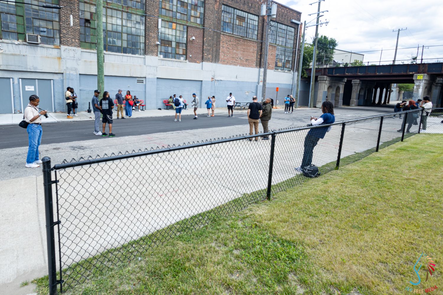 People gathered on an urban basketball court next to a black chain-link fence near an industrial building.