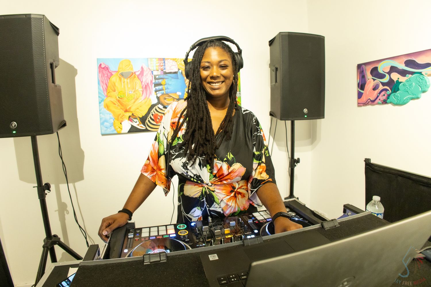 Smiling female DJ with headphones working at a DJ booth with speakers and colorful artwork in background.