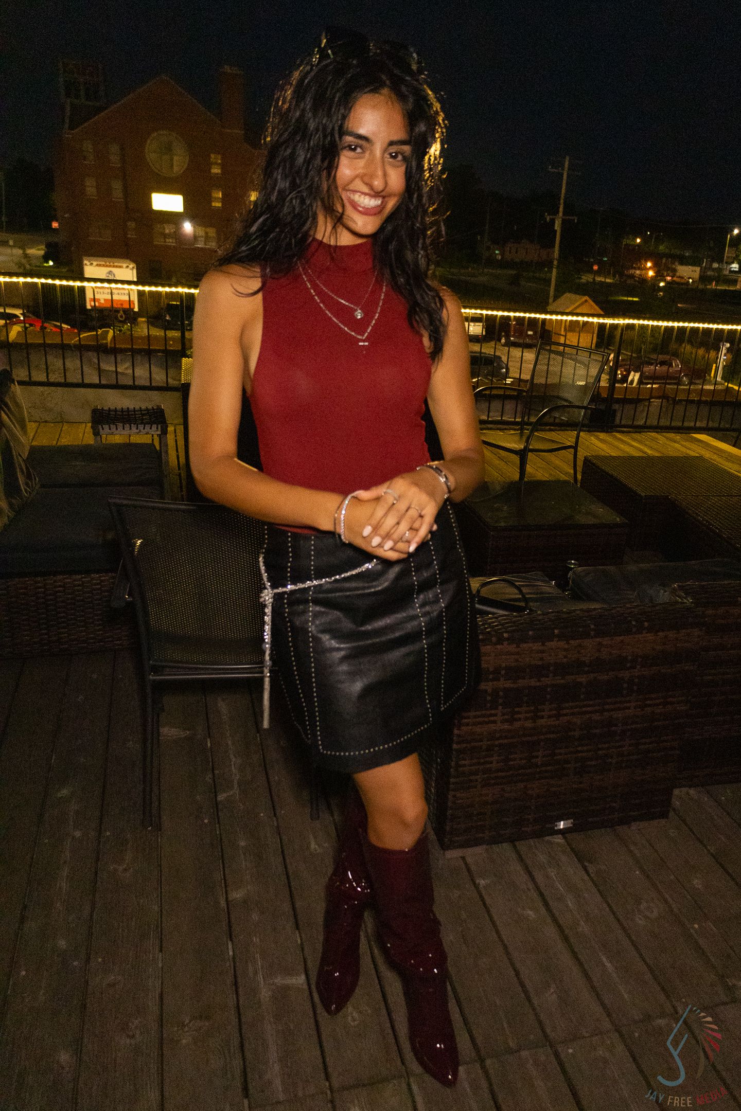 A young woman in a red sleeveless top, black leather mini skirt, and burgundy boots poses on a rooftop patio at night.