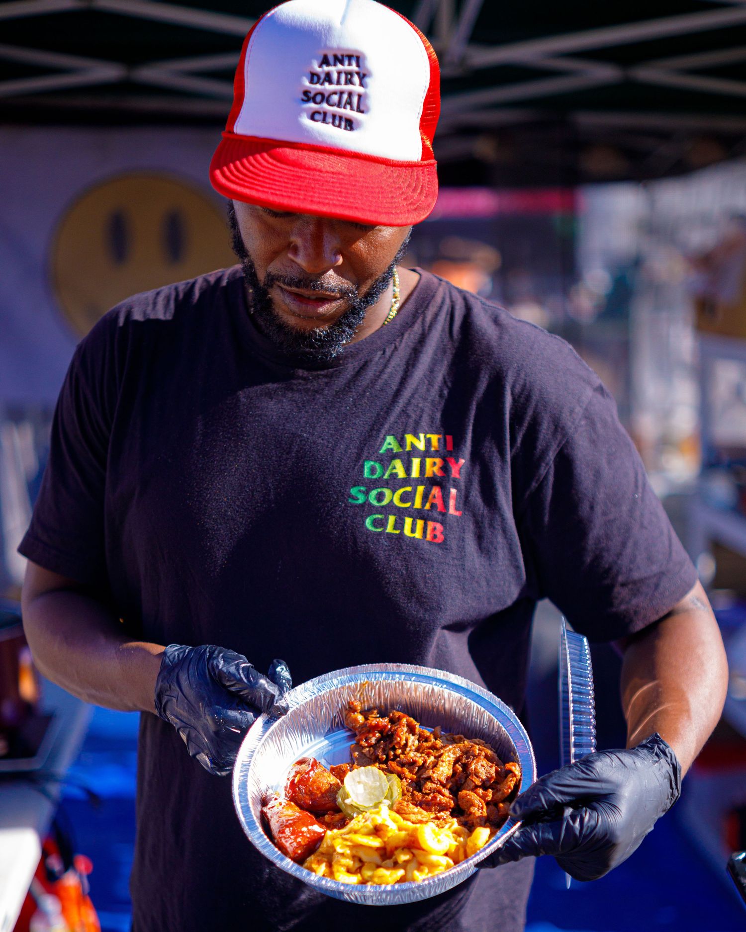 Man in Anti Dairy Social Club shirt holds plate of vegan food at outdoor festival.