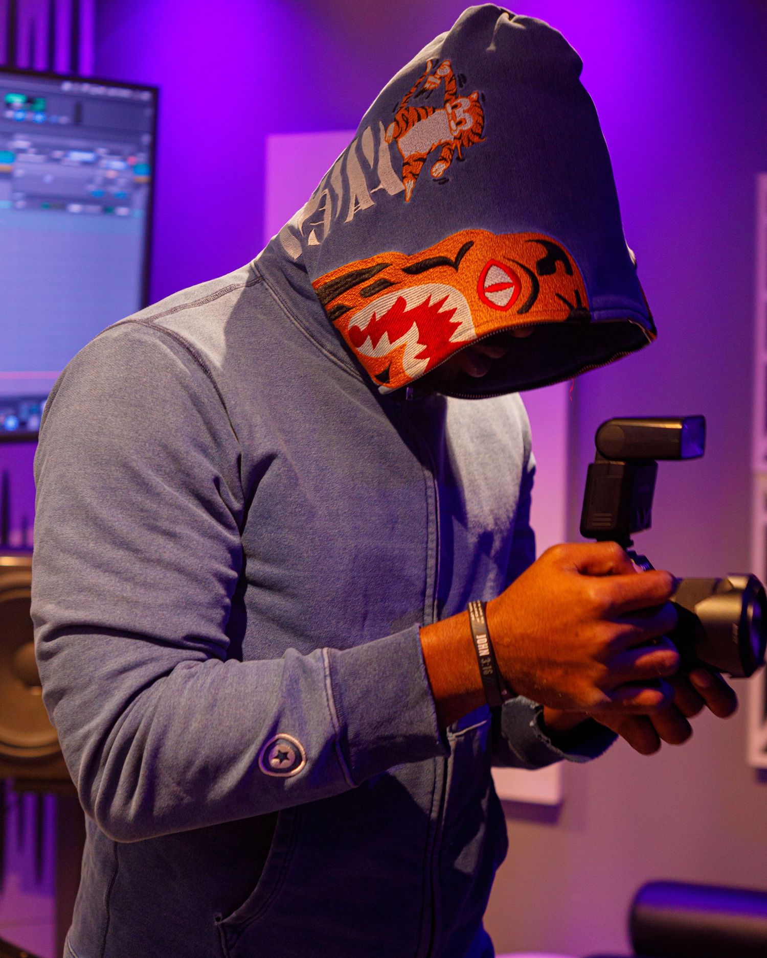 Person in hooded sweatshirt holding camera gimbal in purple-lit studio environment.