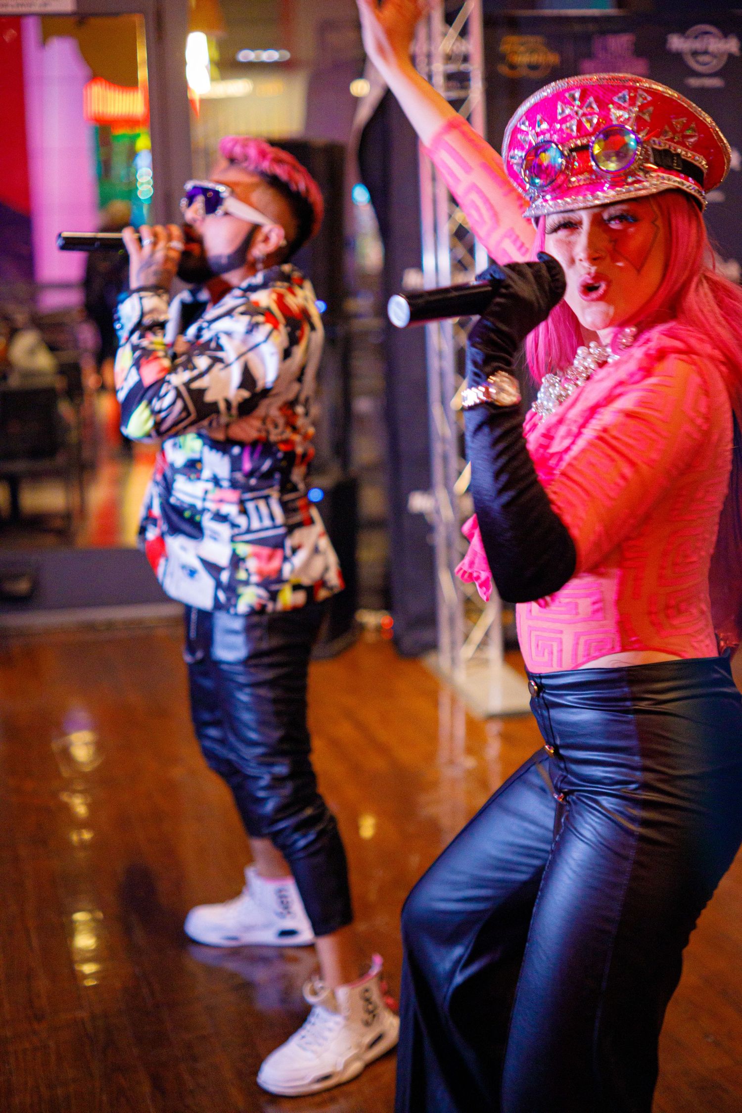 Two performers in pink outfits and decorated hats singing with microphones on stage.