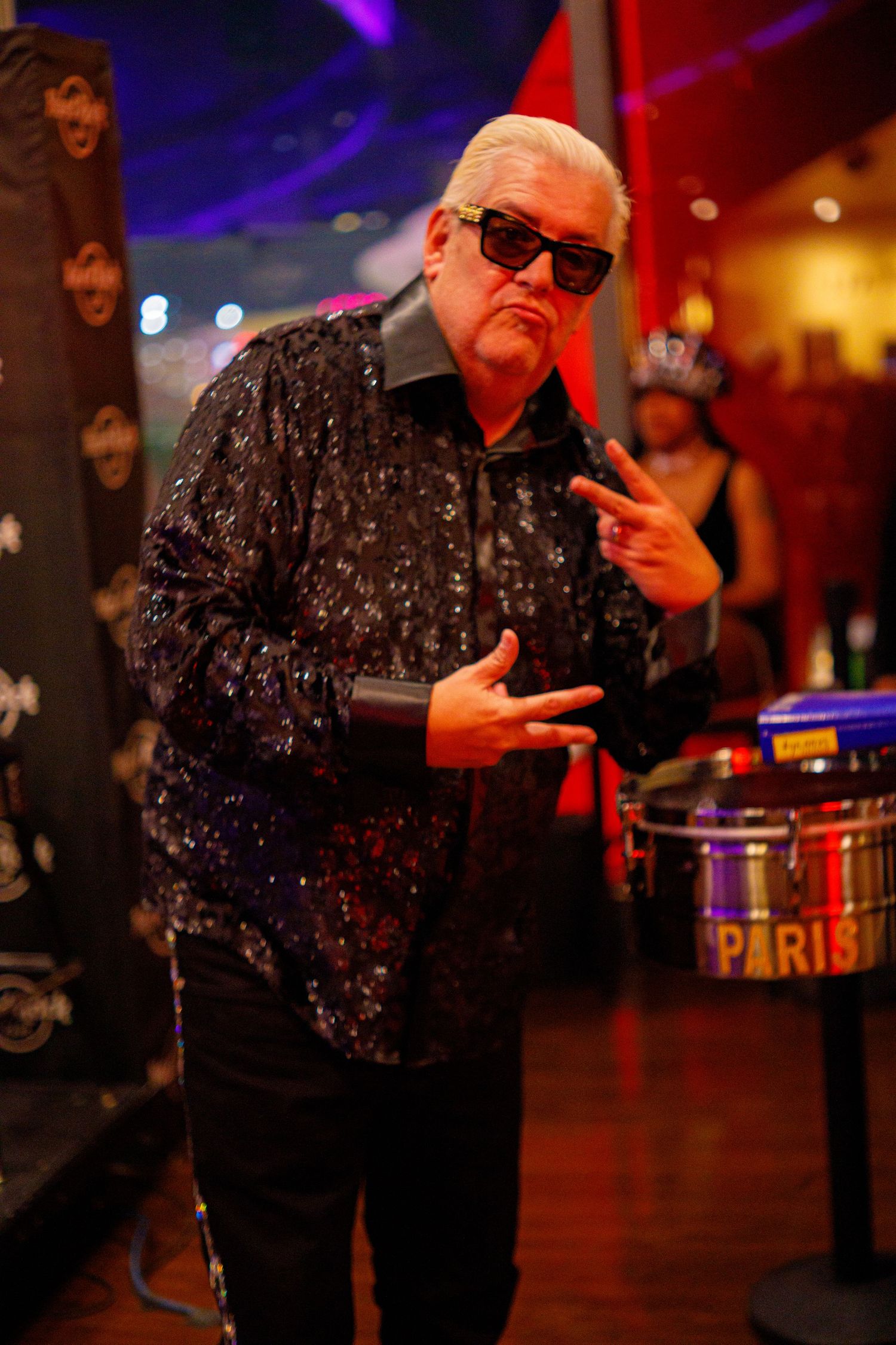 Older man in black sequined shirt and sunglasses poses confidently at a vibrant nightclub event.