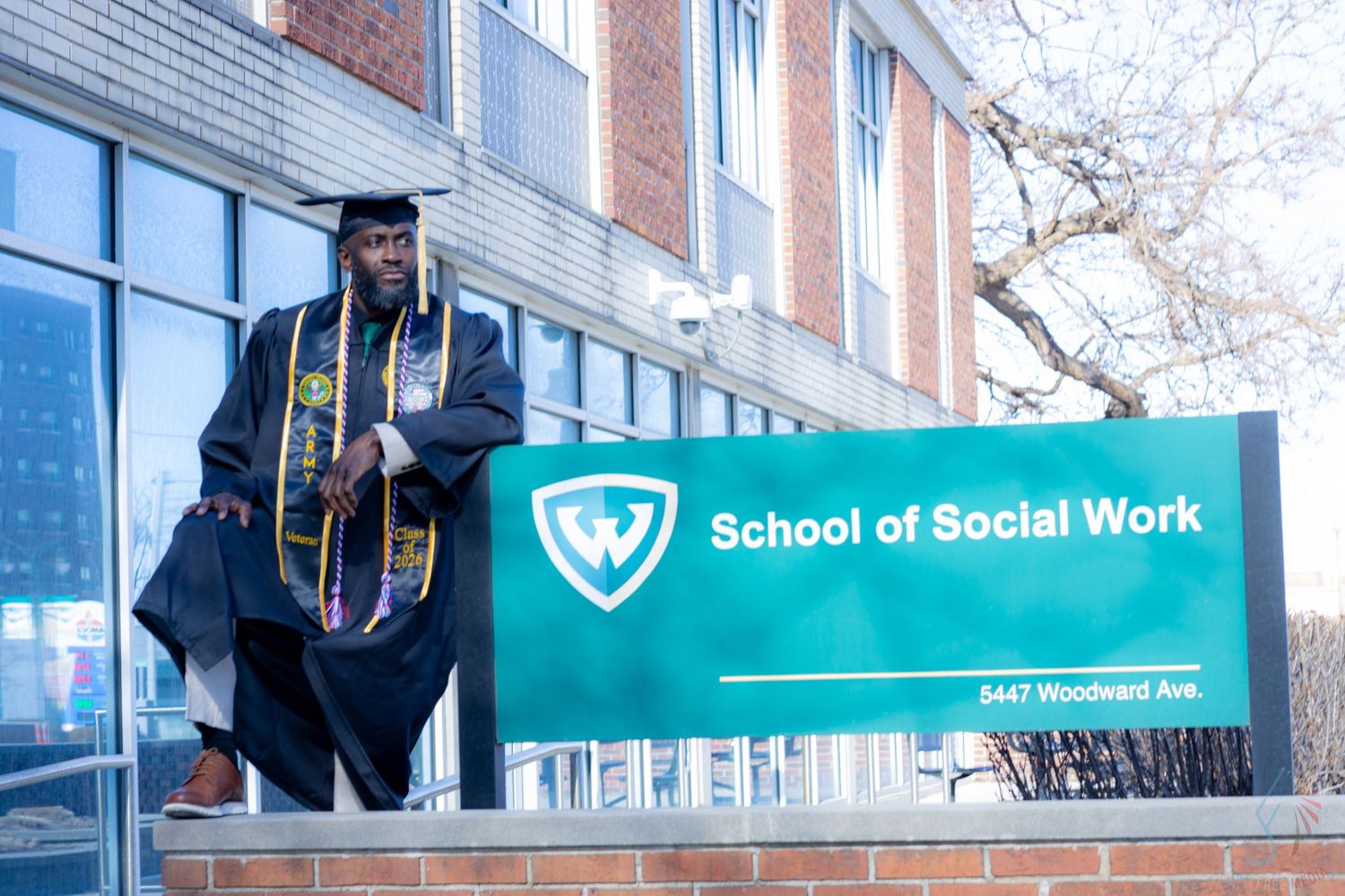 Graduate in cap and gown poses proudly by Wayne State University School of Social Work sign on Woodward Ave.