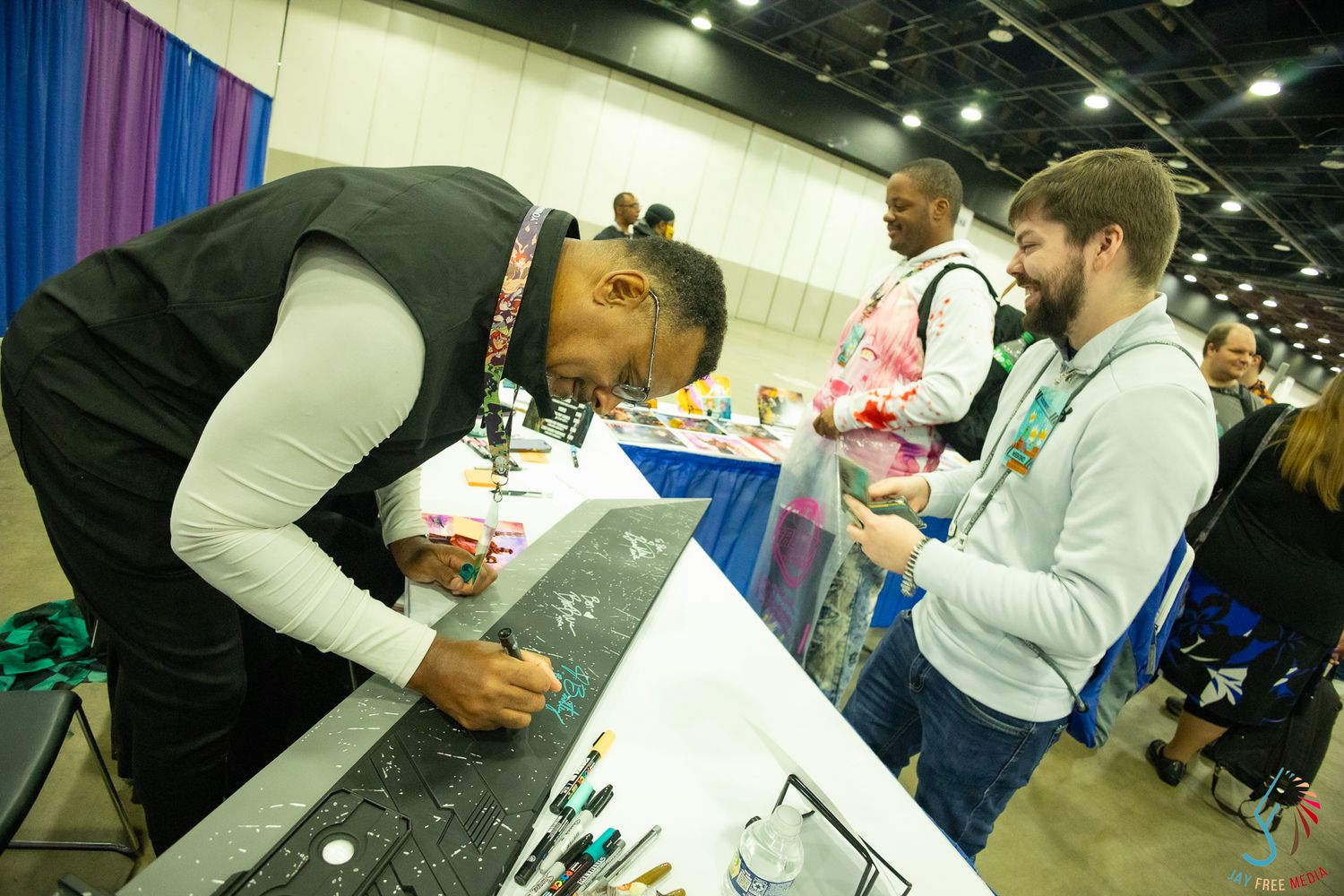 Artist signing autographs at a convention booth while fans wait in line at an indoor exhibition hall.