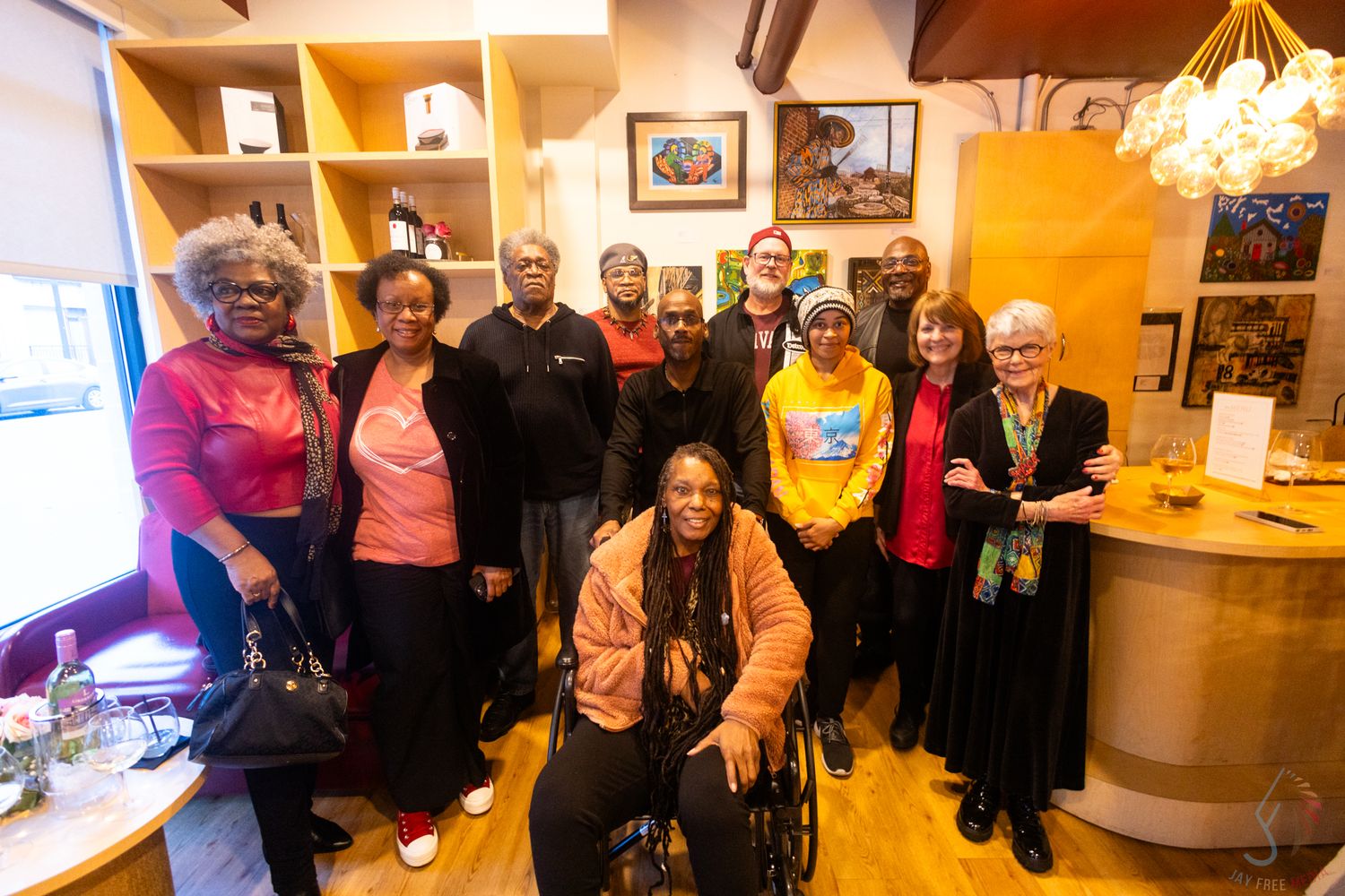 Diverse group of people posing together indoors, including a woman in a wheelchair, smiling in a warmly lit room.