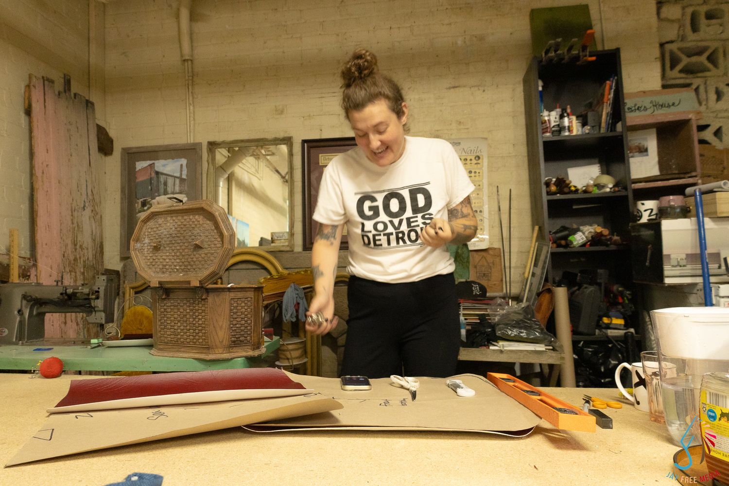 Woman in God Loves Detroit shirt works at a wooden workbench in a Detroit art studio.