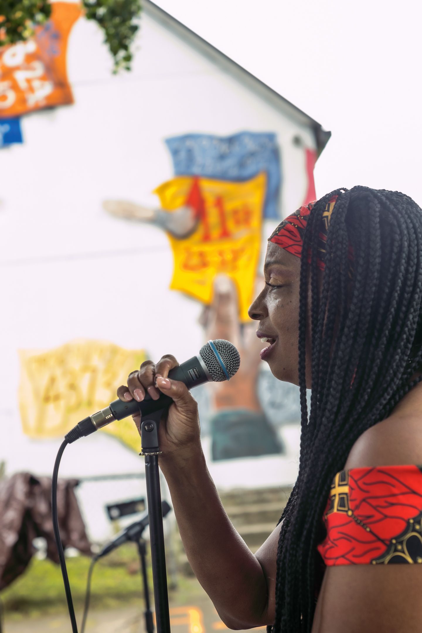 Young Black woman with braids singing into a microphone at an outdoor festival with colorful banners in background.