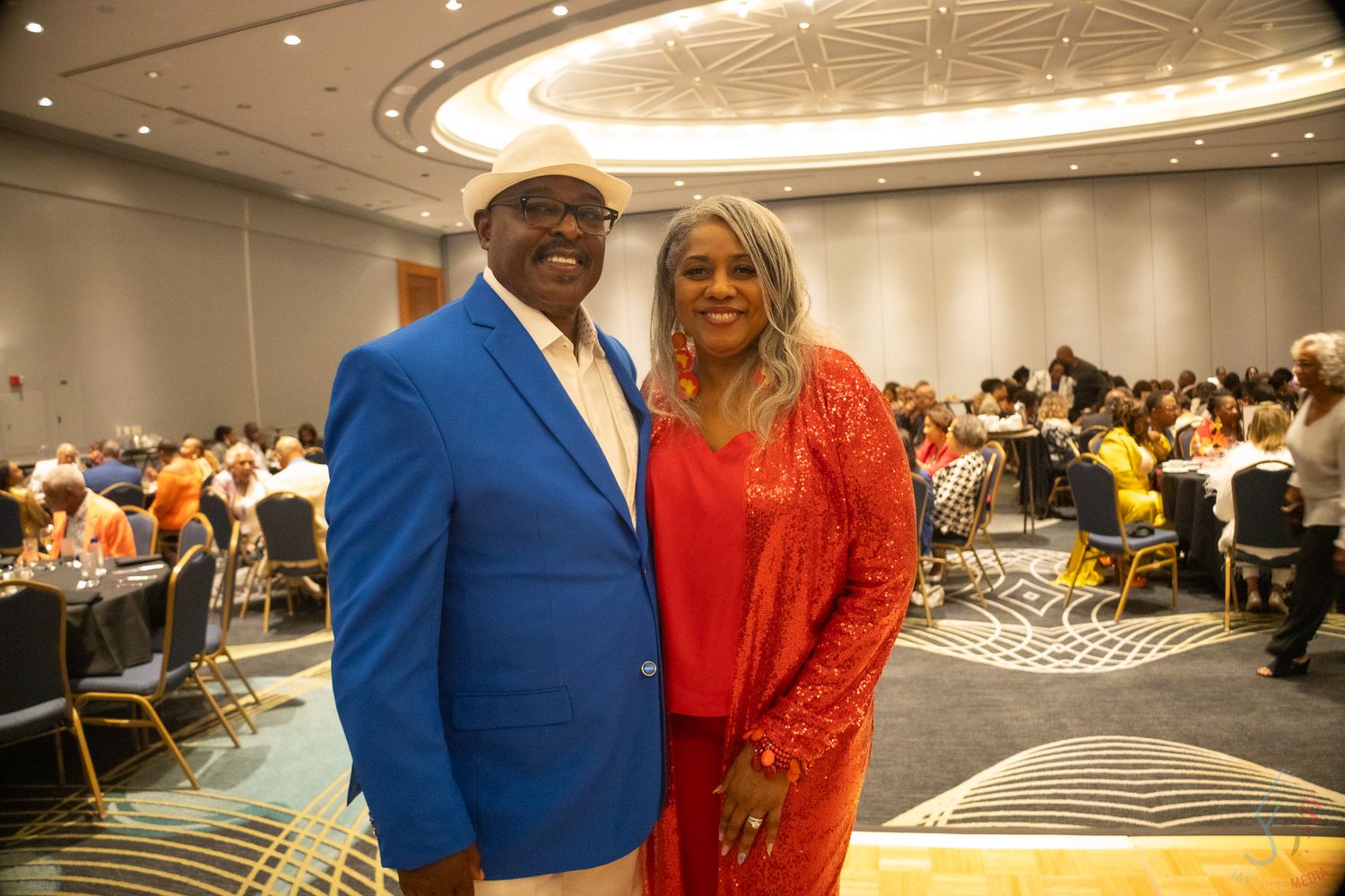 Couple in formal attire, man in blue blazer and woman in red dress, pose at a banquet hall.