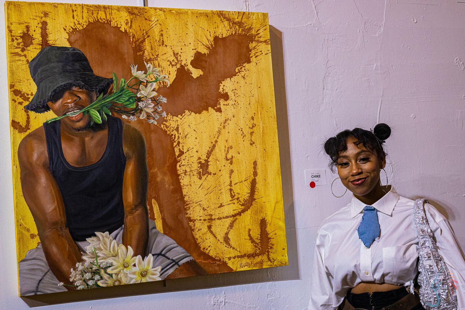 A woman smiles beside a vibrant painting of a figure with flowers at an art gallery exhibition.