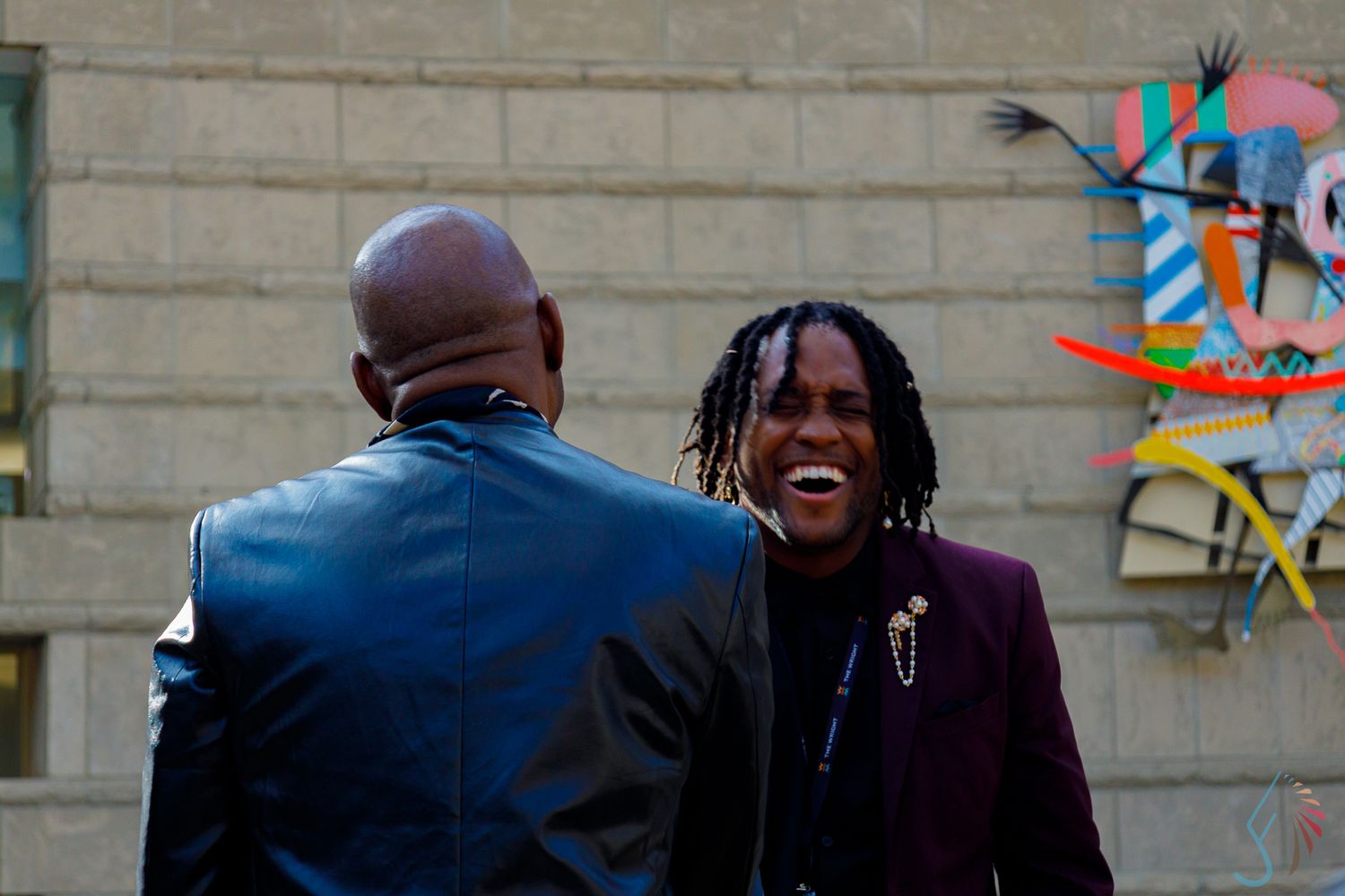 Two men greeting each other outdoors near a colorful bird mural on a stone wall.