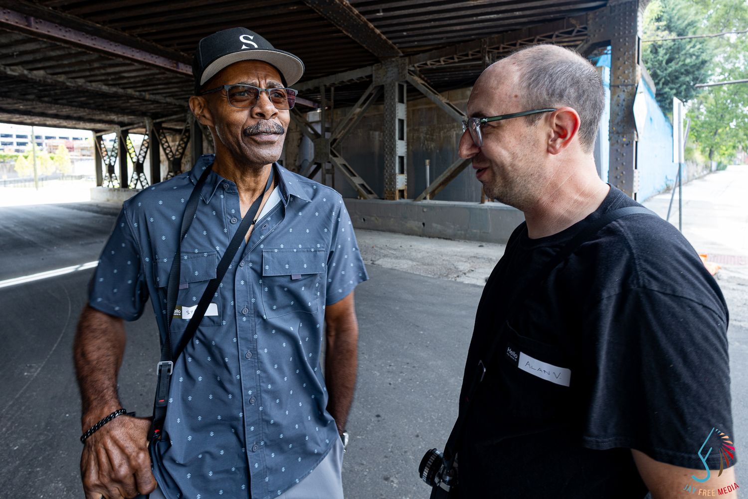 Two men smiling and conversing outdoors under a bridge structure on a sunny day.