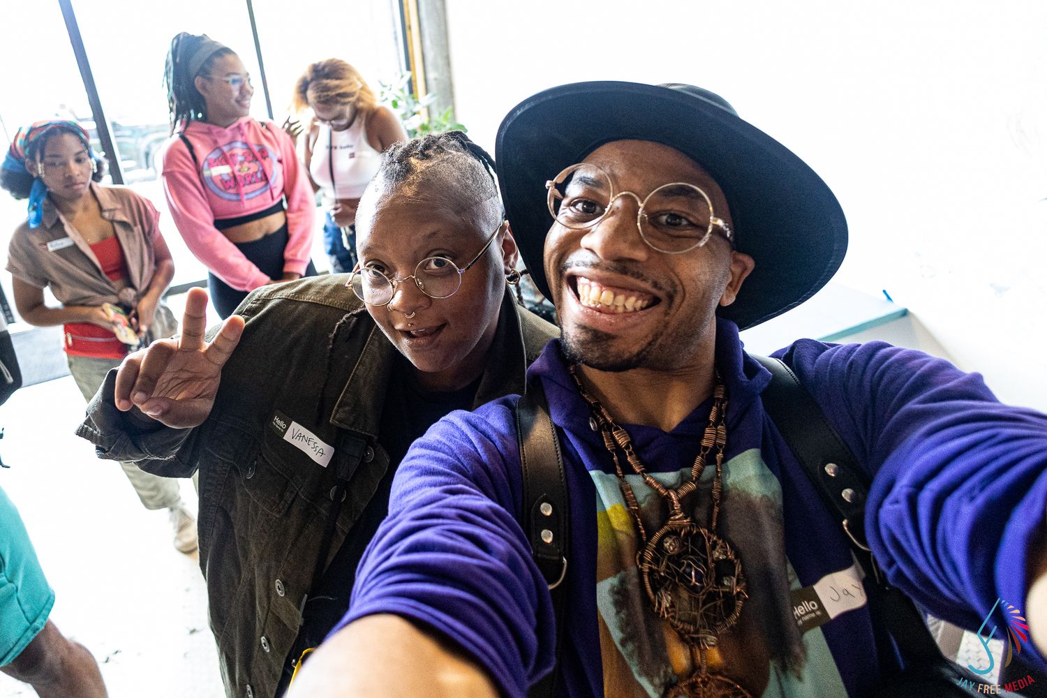 Two friends take a joyful selfie at an indoor event, one flashing a peace sign and the other wearing a stylish hat.