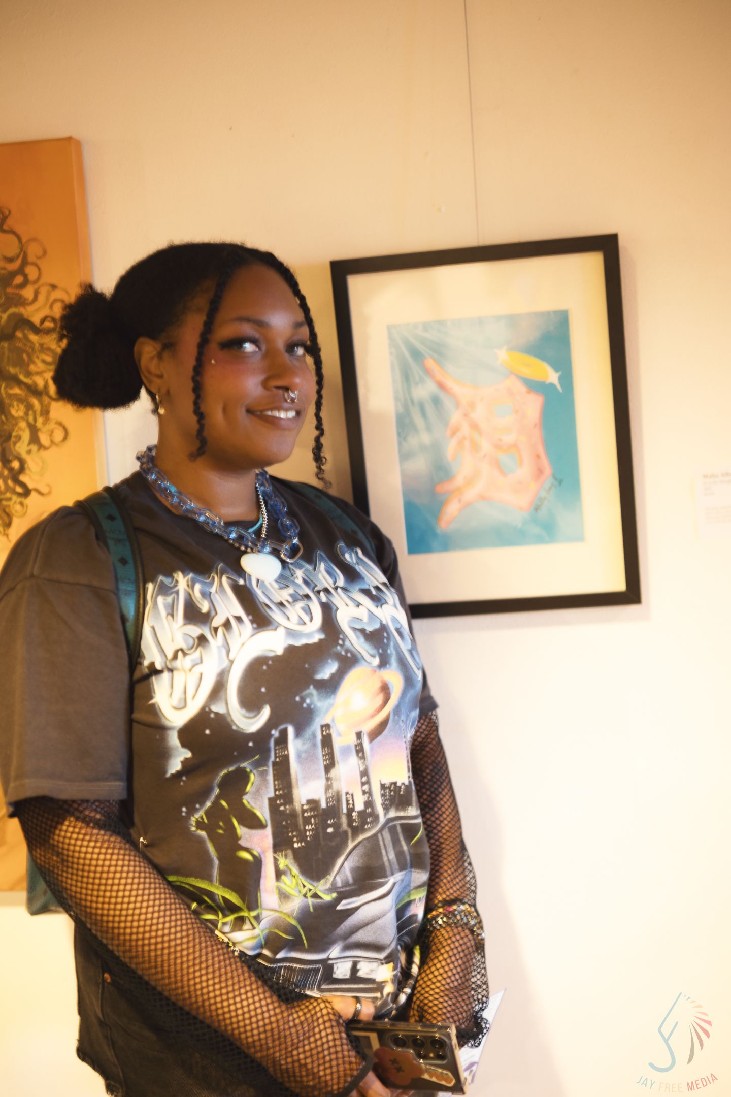Young Black woman smiling in graphic tee and fishnet sleeves standing in front of framed artwork in gallery.