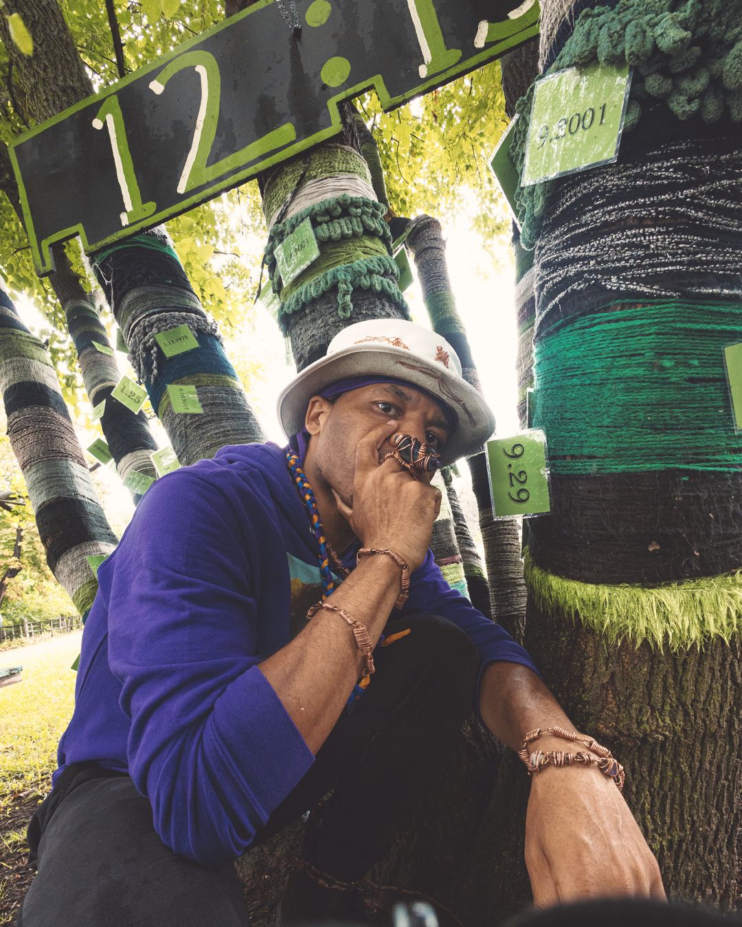 Man in white hat and purple shirt poses thoughtfully among yarn-bombed trees with green tags in a park.