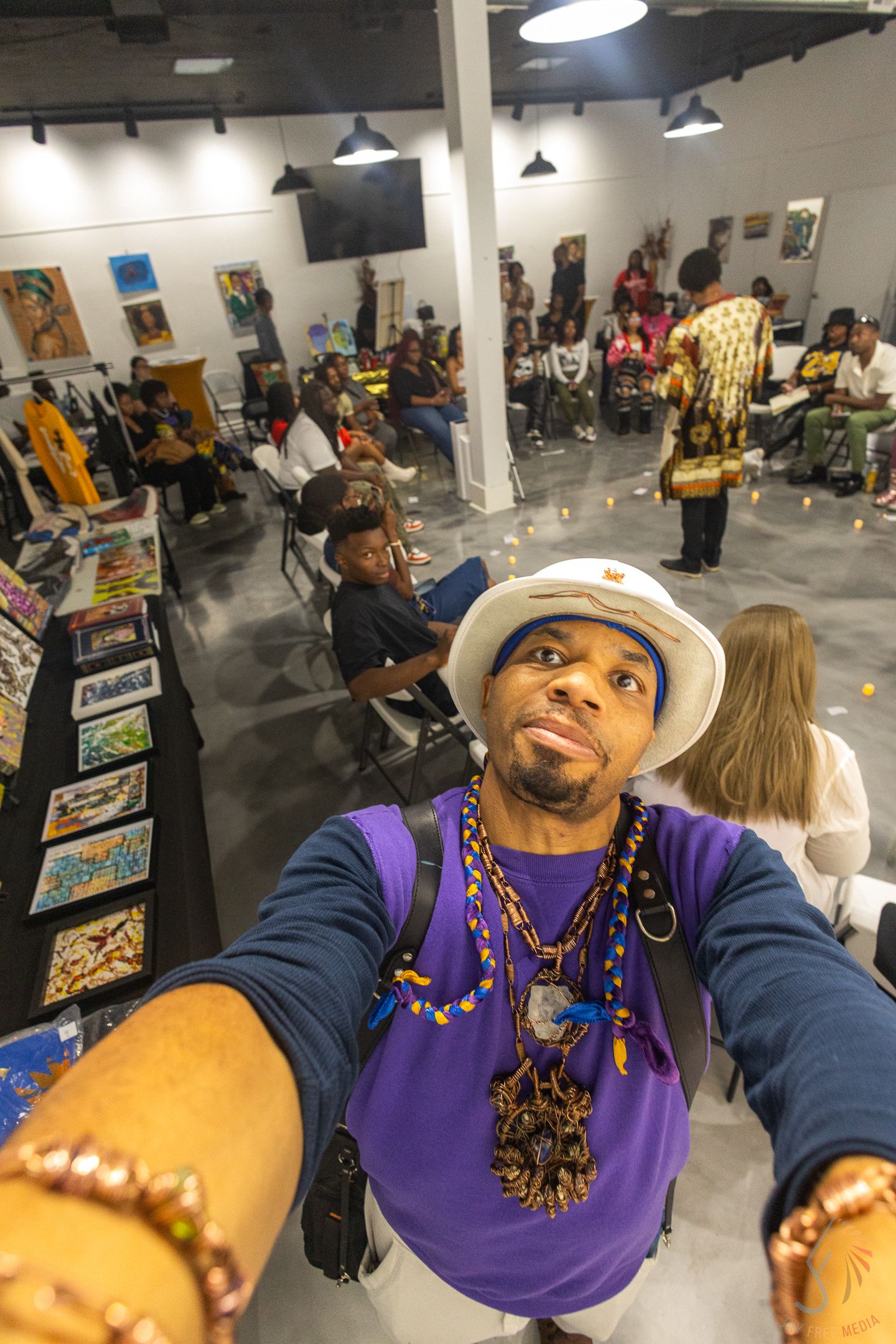 A man in a white hat and purple shirt takes a selfie at a vibrant art gallery event.