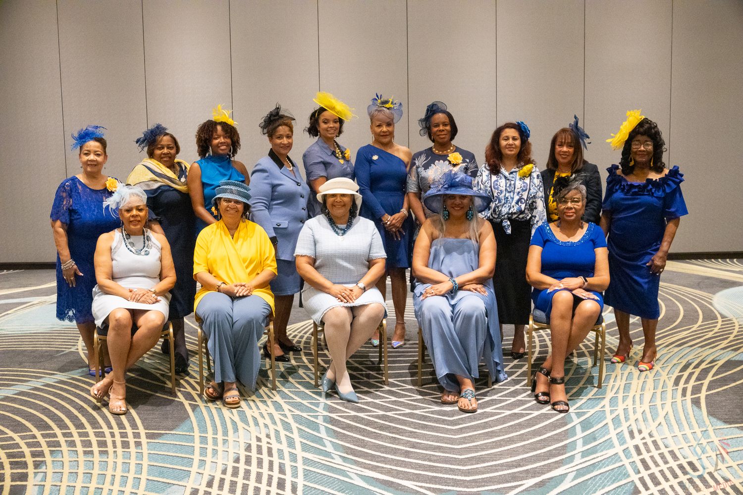 Group of women in blue and yellow outfits and decorative hats posing for a formal group photo indoors.