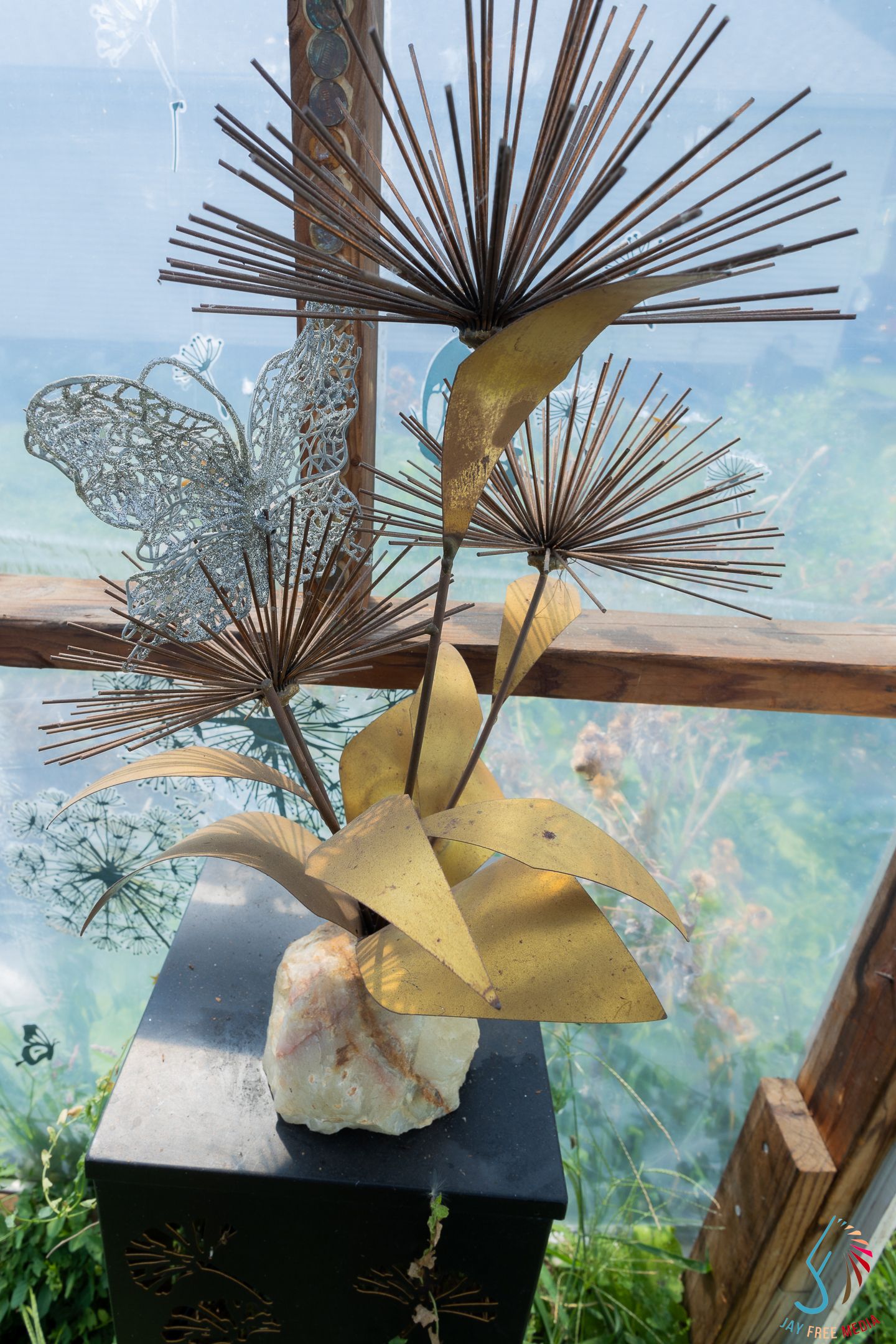 Gold metal sculpture with butterfly and starburst flowers on crystal base, displayed near a window with green garden backdrop.
