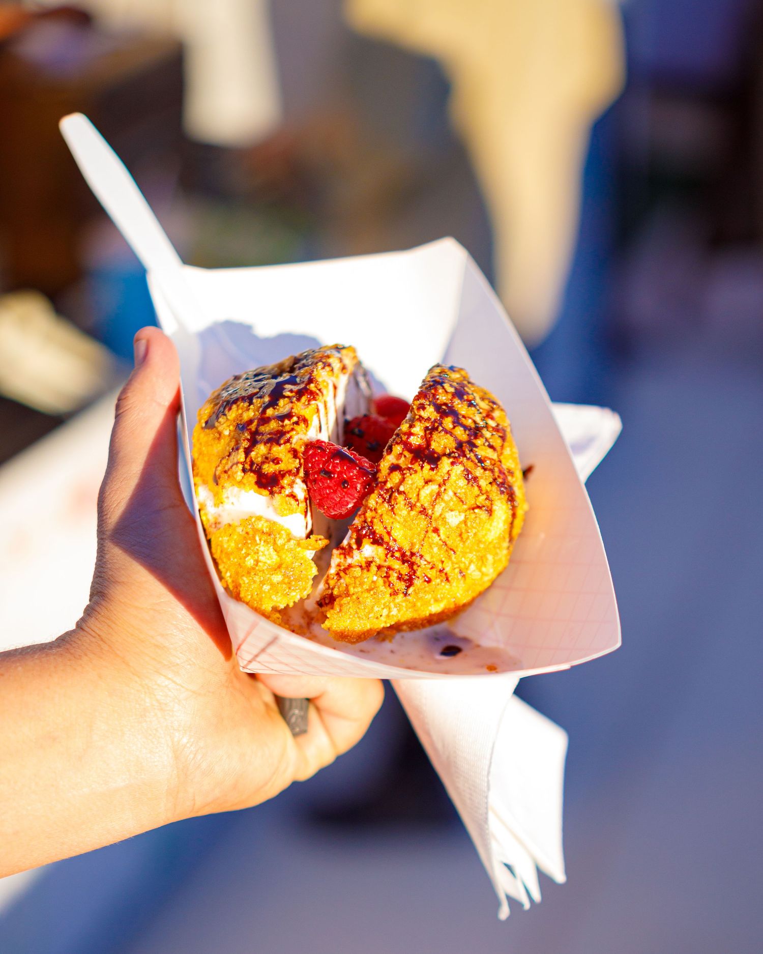 Hand holding a deep-fried dessert with red filling in a white paper tray outdoors.