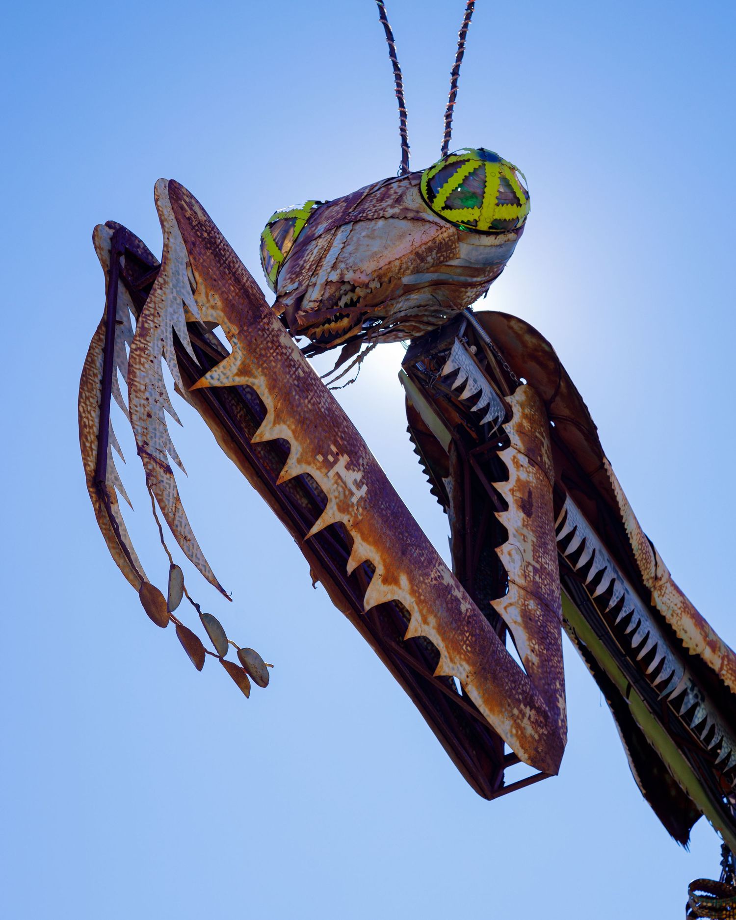 Close-up of a large metal praying mantis sculpture with green eyes against a blue sky.