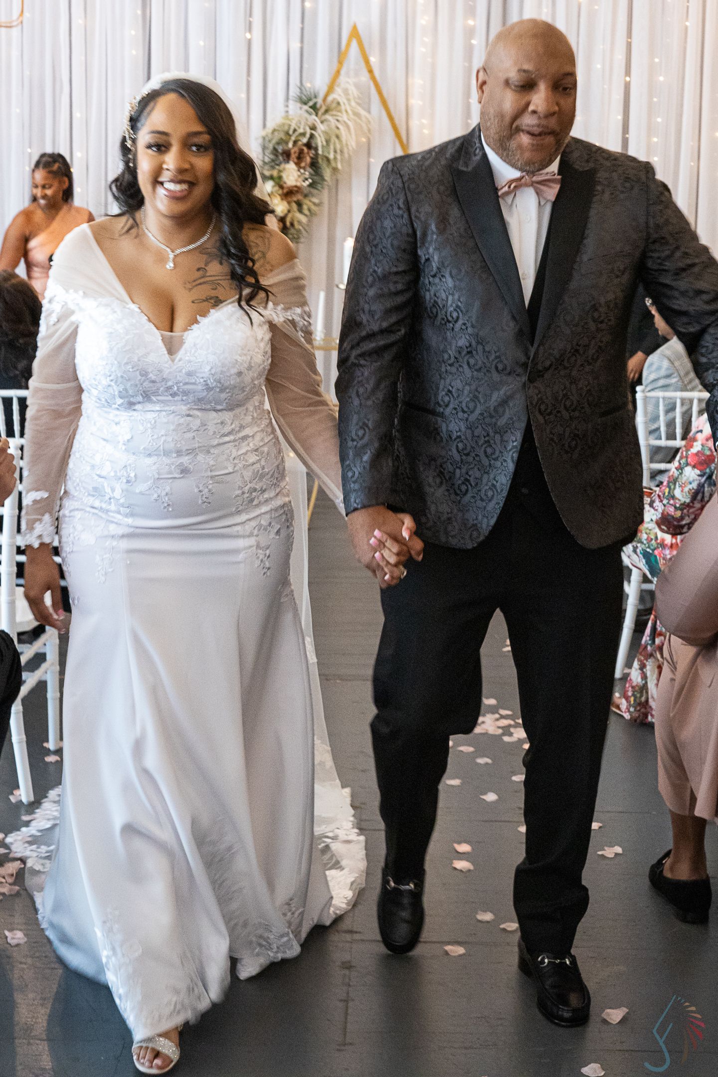 Bride in white lace gown holds hands with groom in black patterned blazer walking down wedding aisle.