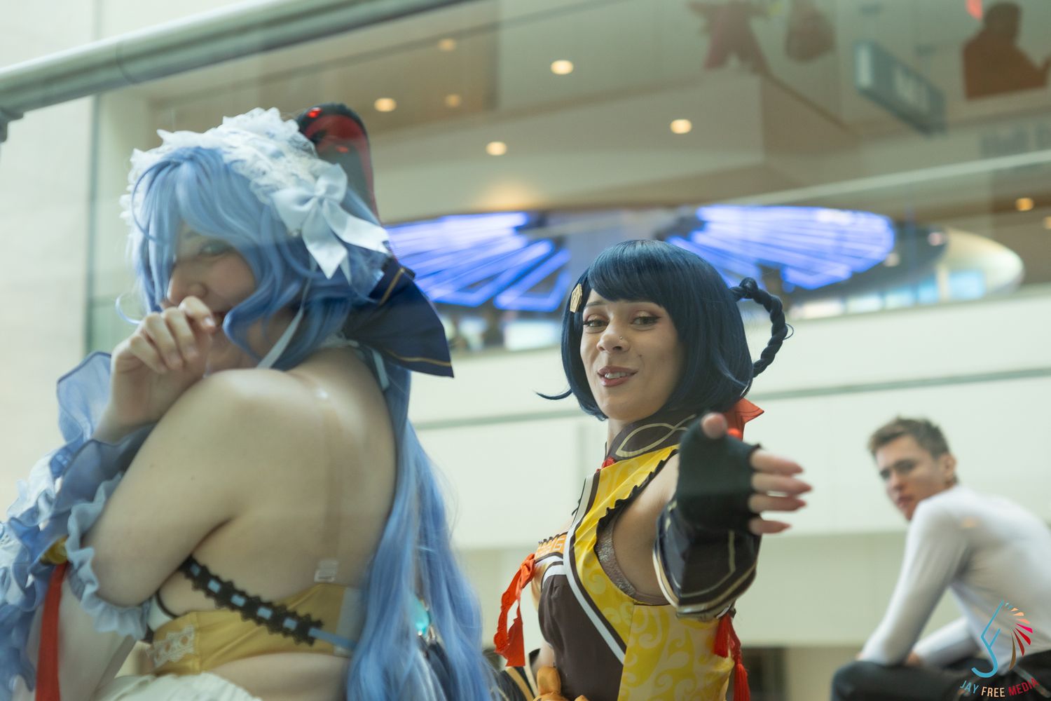 Two anime cosplayers in blue and gold costumes interact dramatically at an indoor convention.
