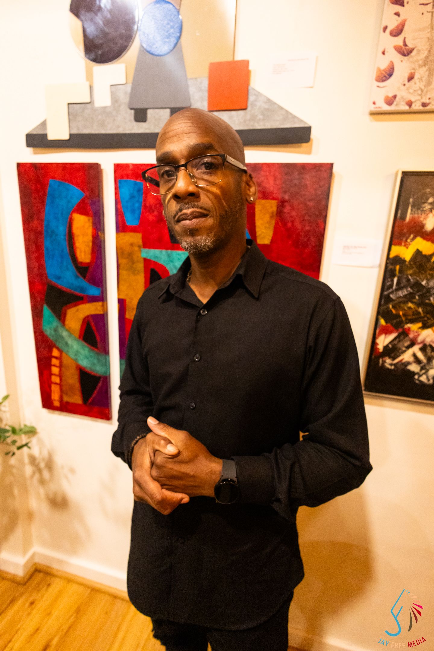 Man in black shirt stands before colorful abstract art paintings in a gallery.