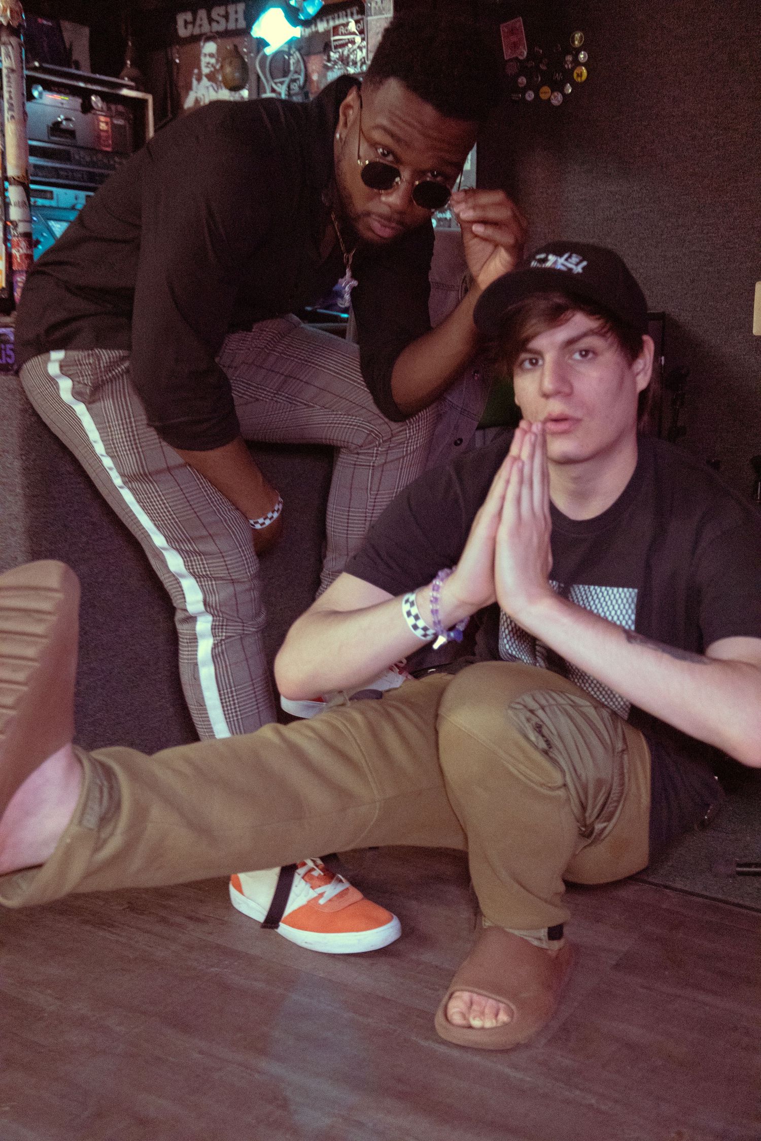 Two young men pose playfully indoors, one standing in plaid pants and sunglasses, the other sitting with hands pressed together.