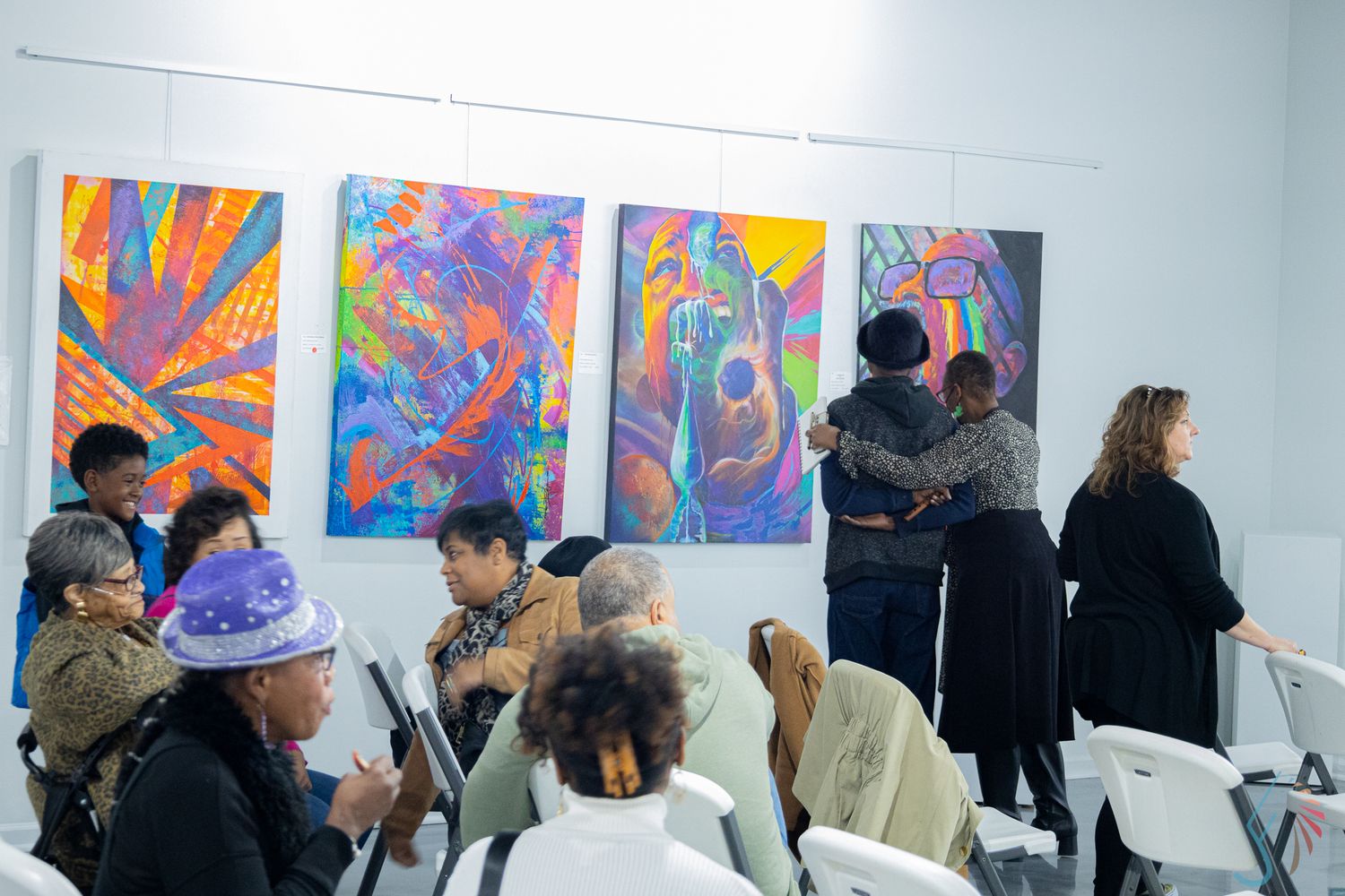 Visitors viewing vibrant colorful paintings at an art gallery exhibition with people seated and standing.