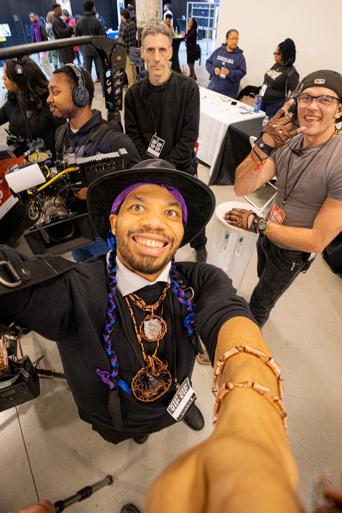 Man in black hat and purple beads takes selfie at indoor event with crew and equipment behind him.