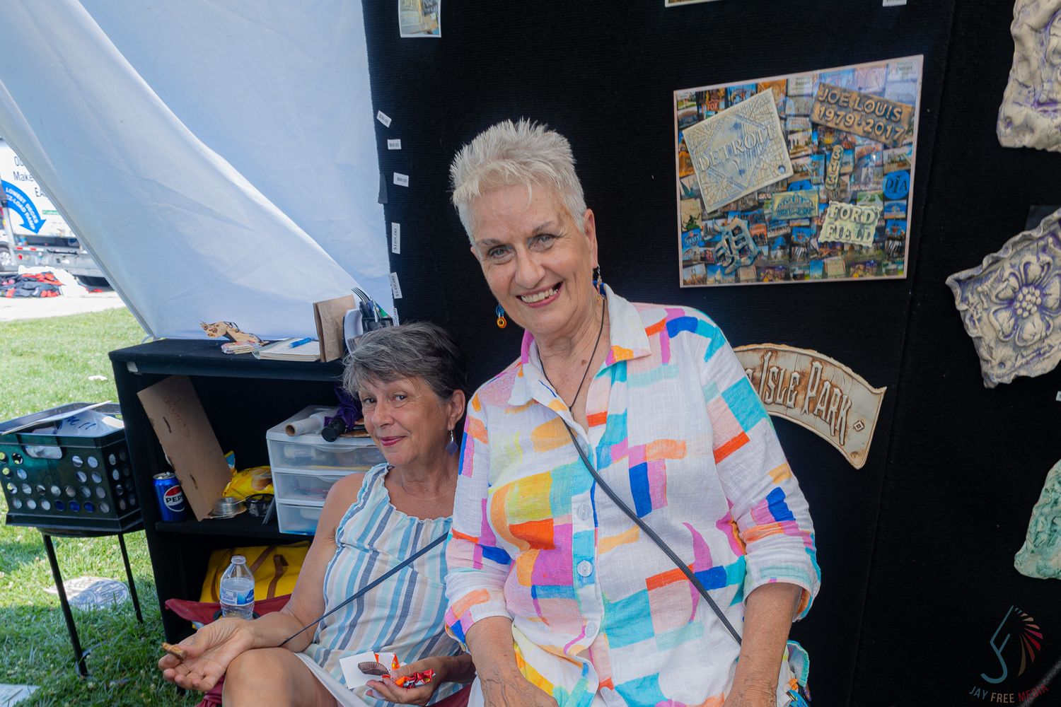 Two smiling women posing together in an outdoor art booth with colorful artwork displayed behind them.