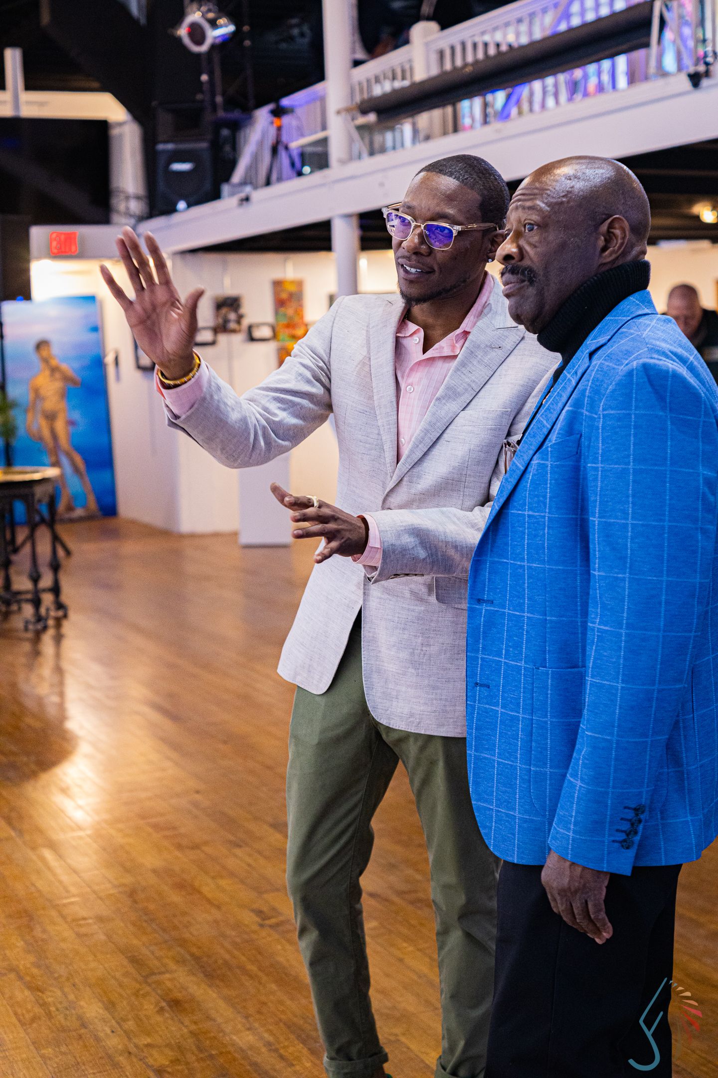 Two well-dressed men in blazers conversing at an art gallery event on a hardwood floor.