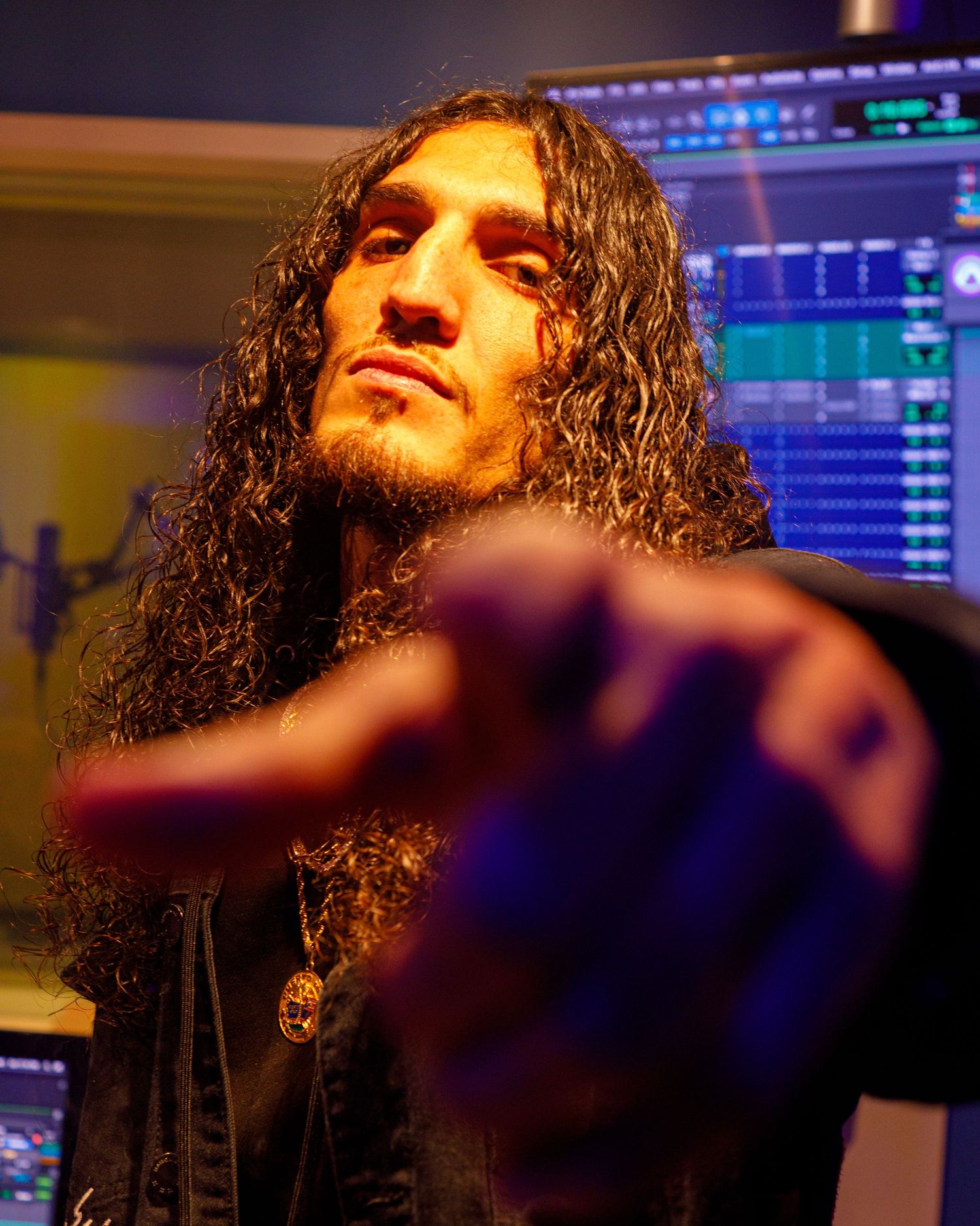 Long-haired rock musician pointing at camera in recording studio with colorful mixing board background.