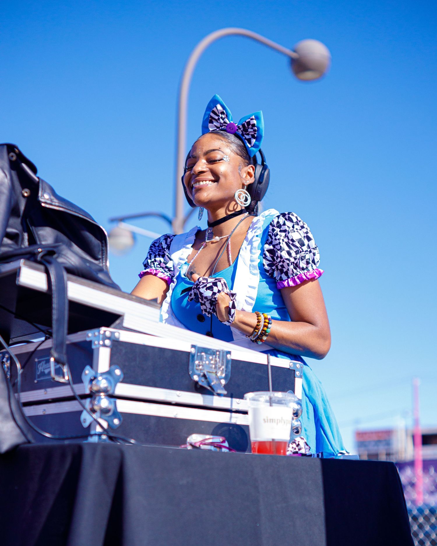 Smiling female DJ in blue outfit with purple bow performs outdoors under clear blue sky.