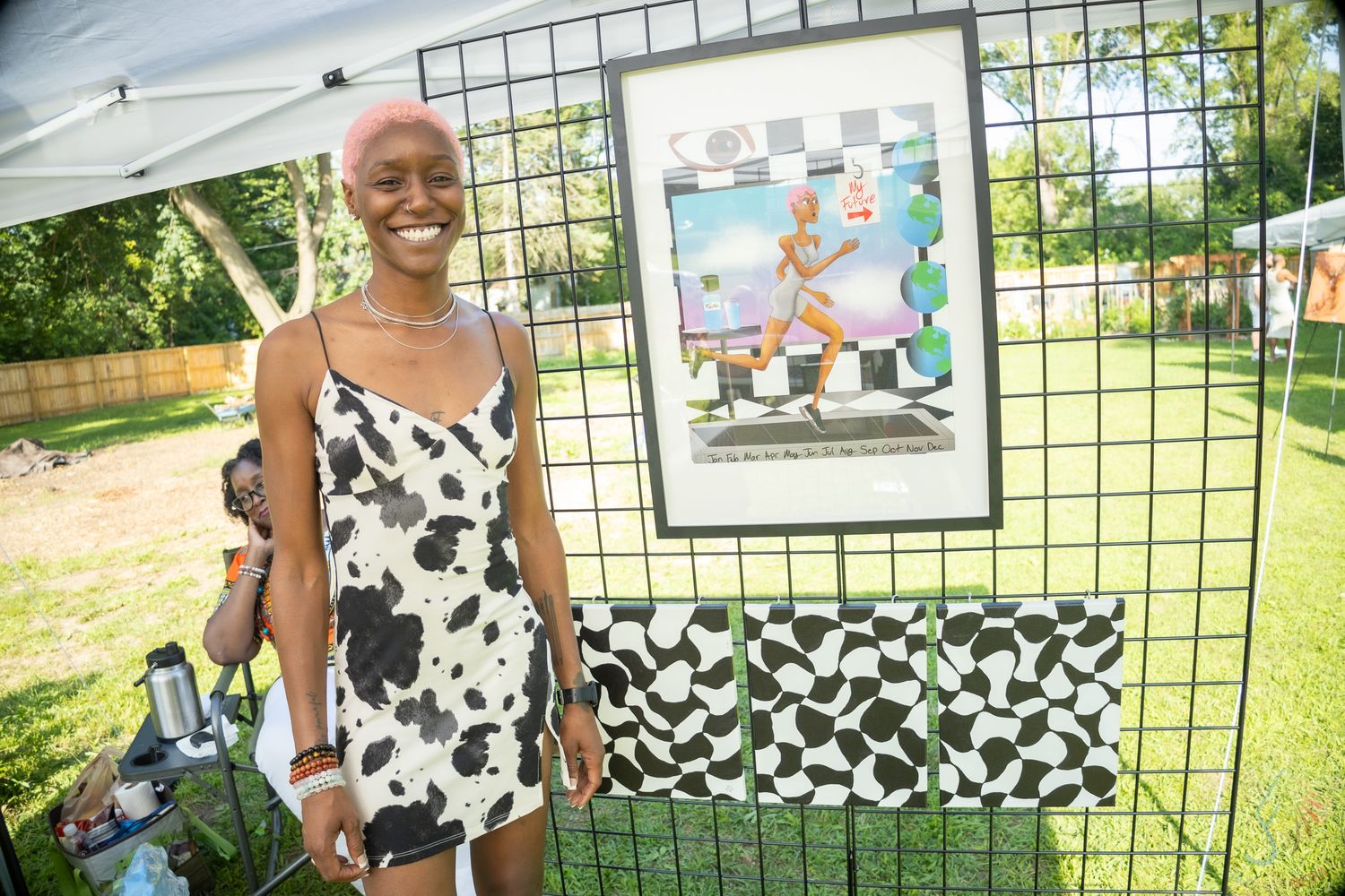 Young woman in cow-print dress smiling beside framed artwork display at outdoor art fair.
