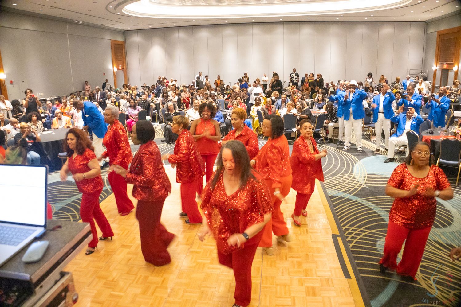 Women in red outfits perform a coordinated dance routine at a large banquet hall event.
