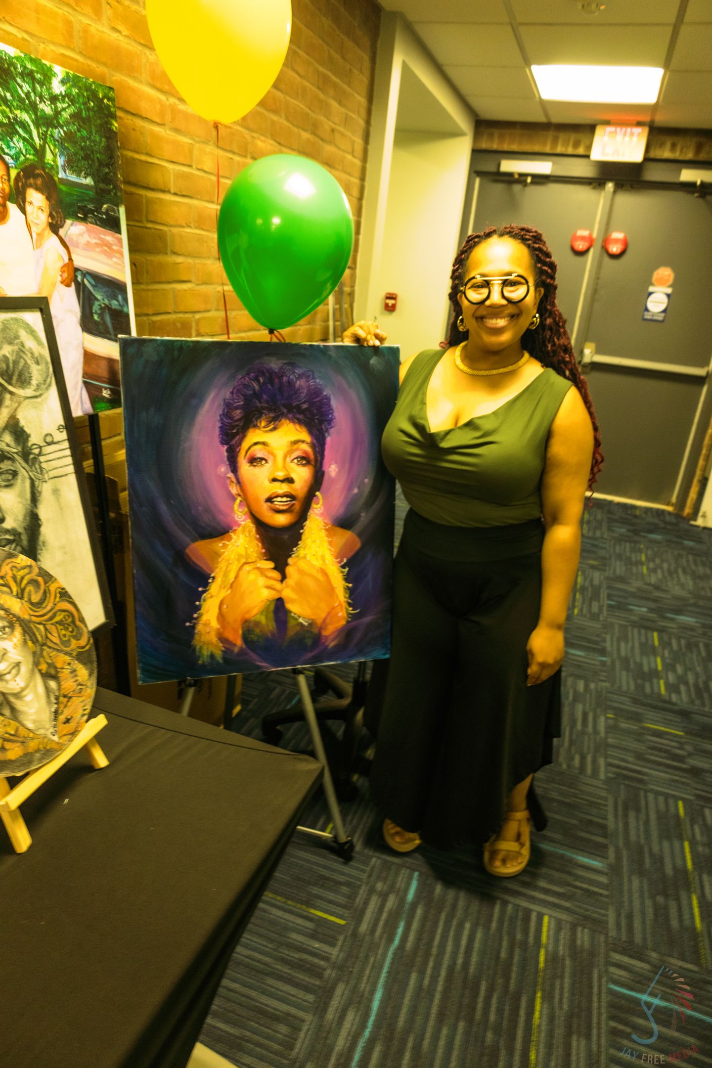 Woman smiling beside a vibrant portrait painting on an easel, with green and yellow balloons at an art exhibition.