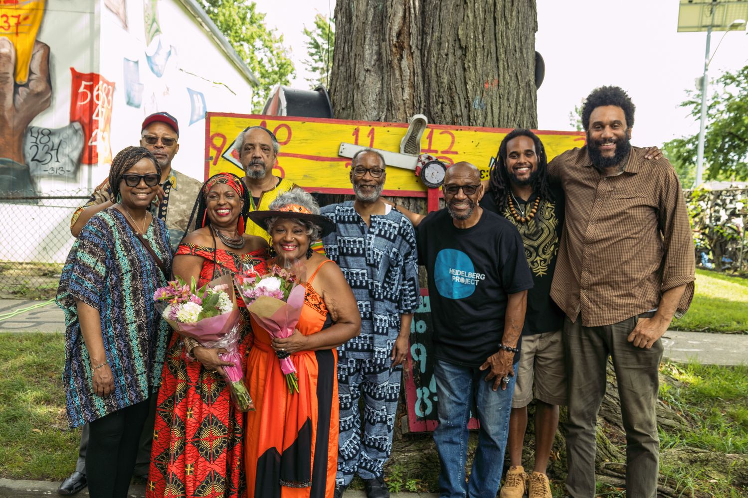 A diverse group of people posing together outdoors near a colorful mural and tree in a vibrant community setting.