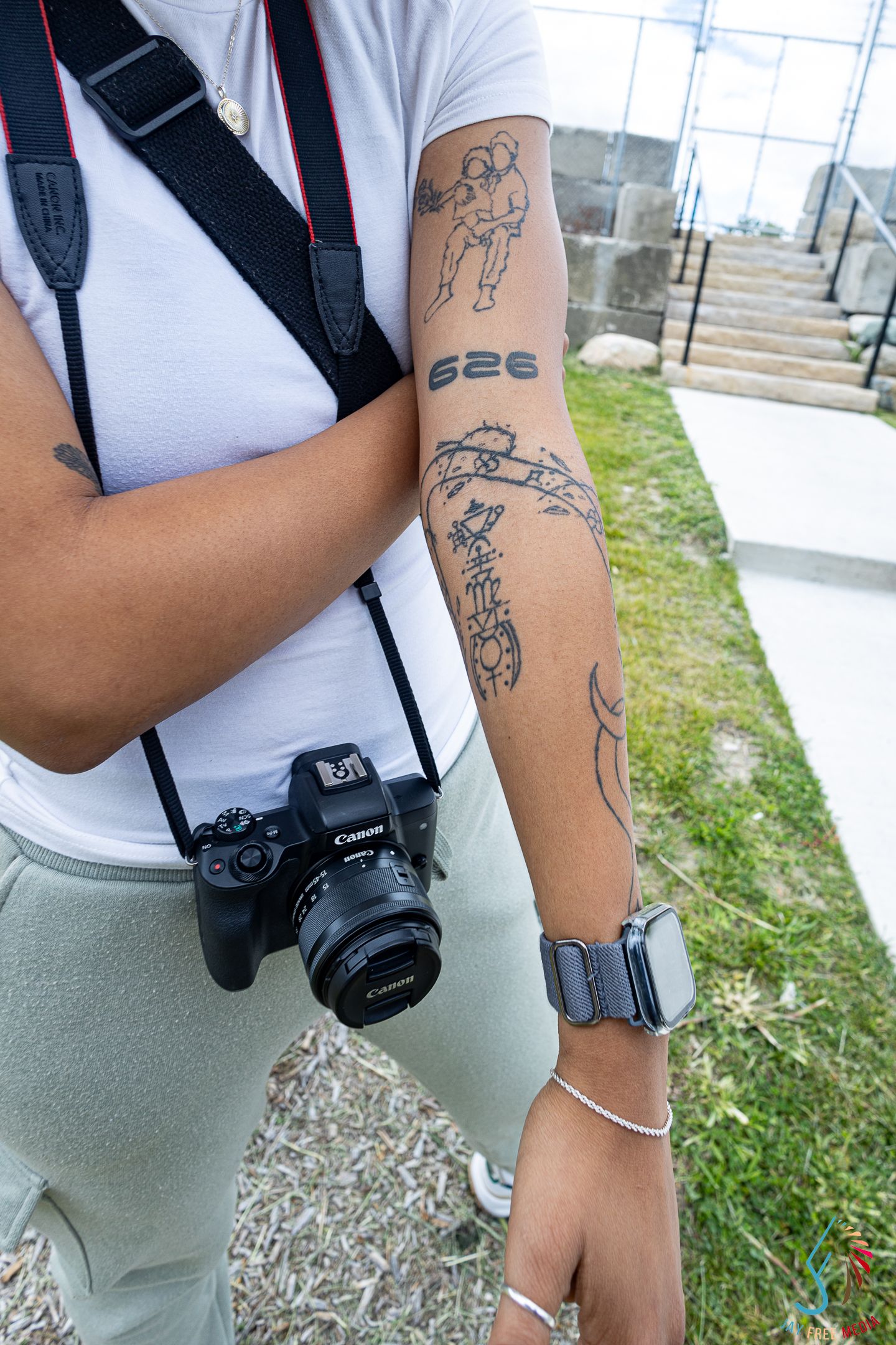Close-up of tattooed arm with 626 ink holding Canon camera with strap outdoors.