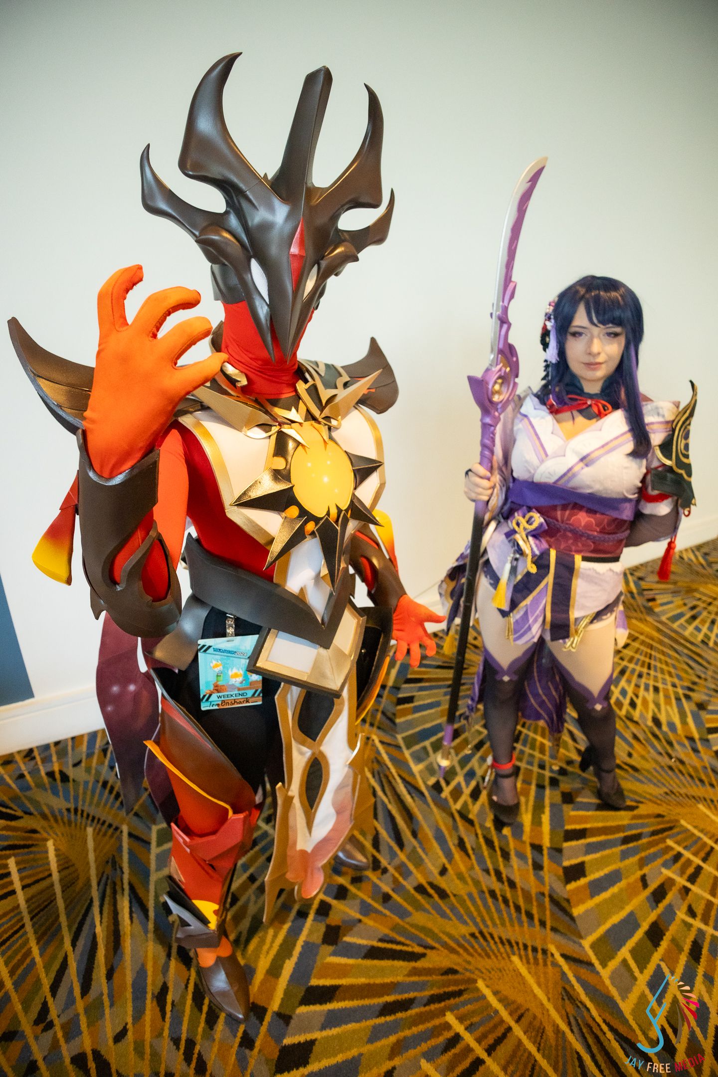 Two cosplayers in elaborate fantasy armor costumes pose at an anime convention, one holding a bow.