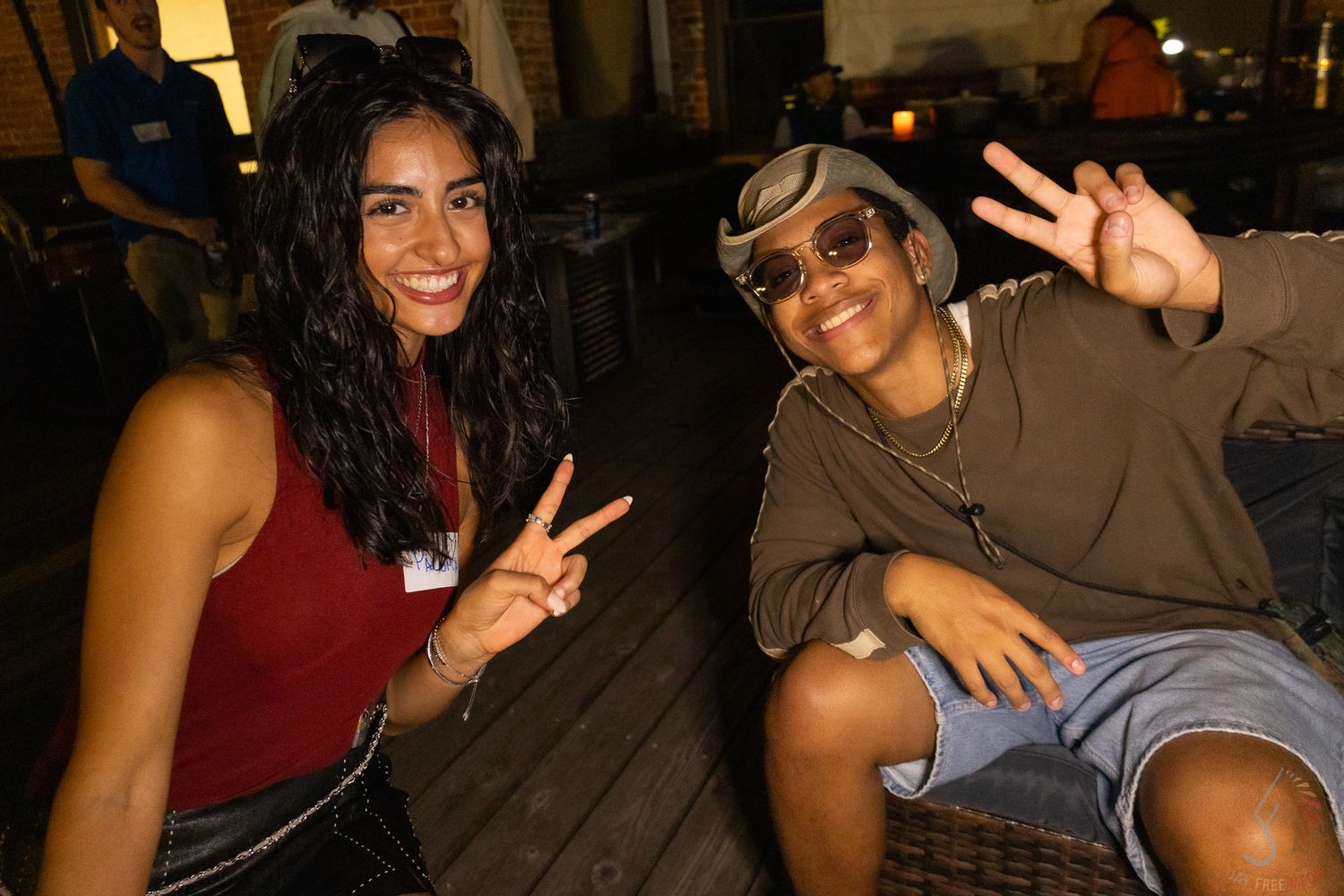 Two young women smiling and making peace signs while seated at an outdoor evening event.