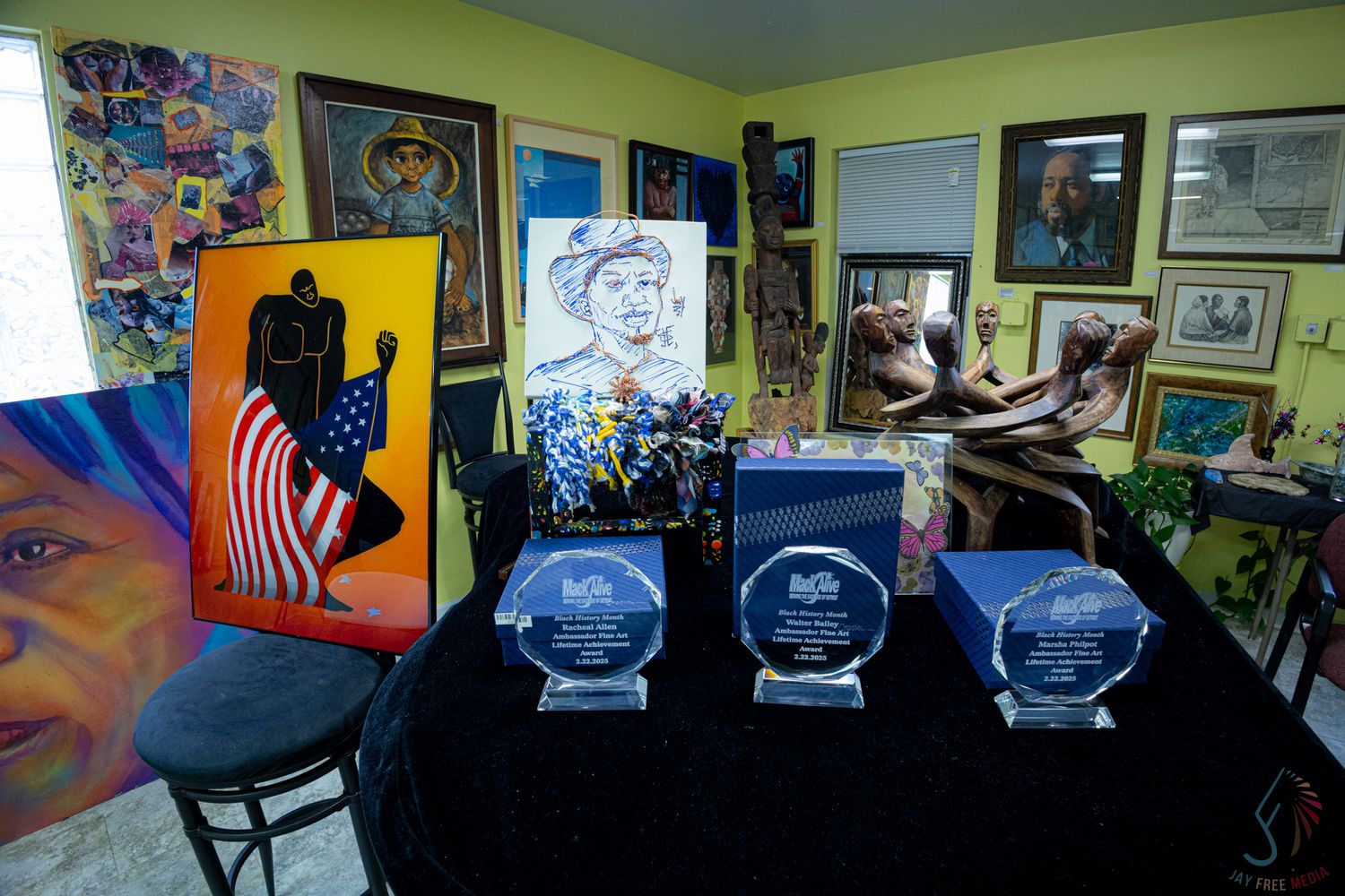 Art studio display with crystal awards, colorful paintings including American flag artwork, sculptures, and framed pieces on yellow walls.