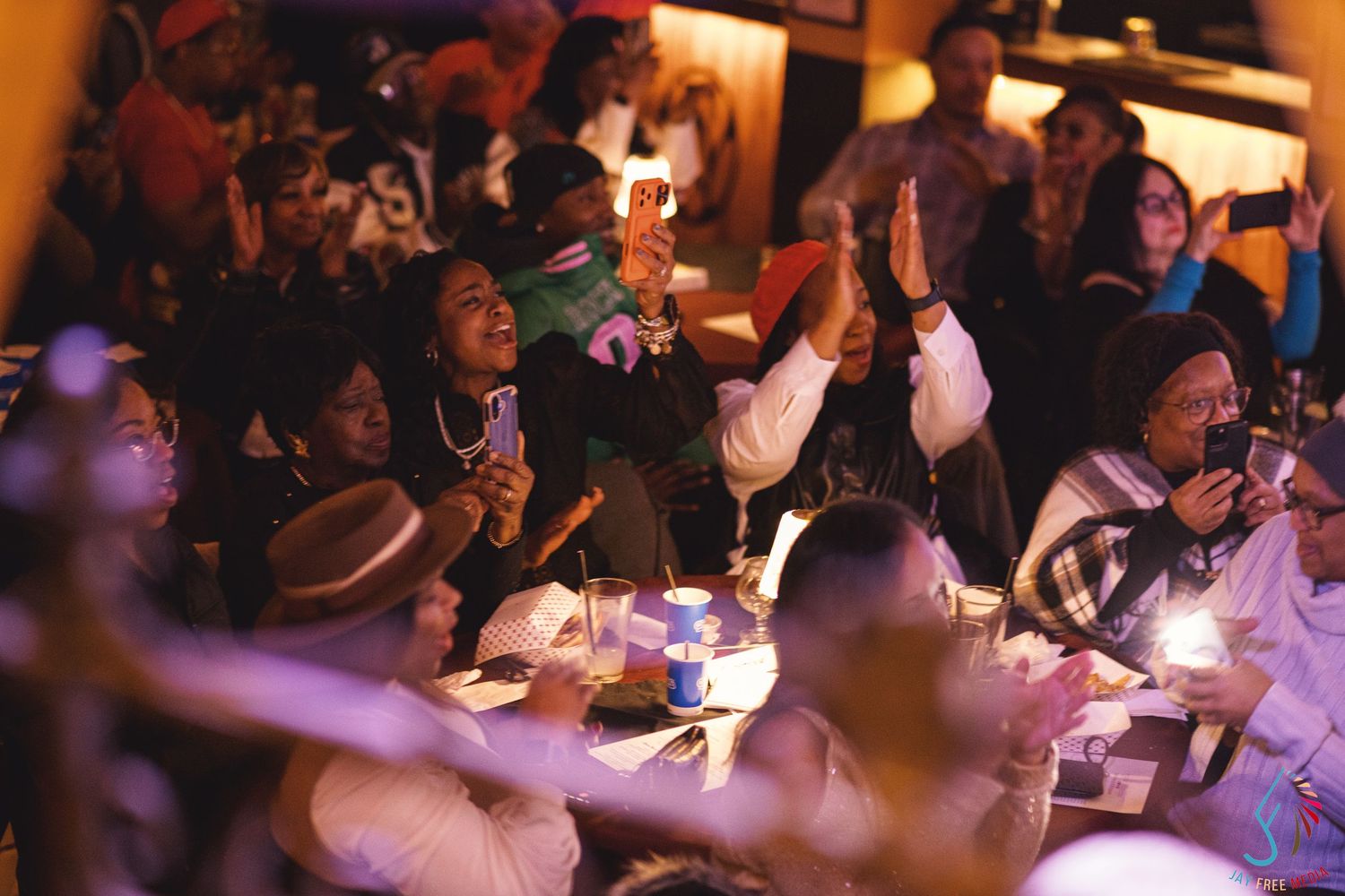 Diverse crowd of excited people celebrating at a dimly lit restaurant event, raising hands and smiling.