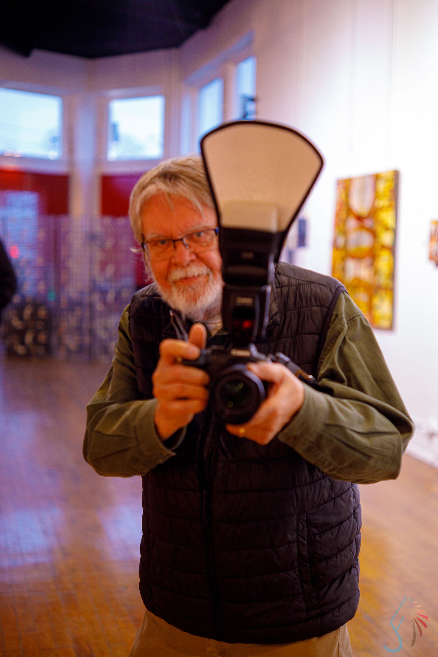 Smiling bearded man holding DSLR camera with flash in art gallery.