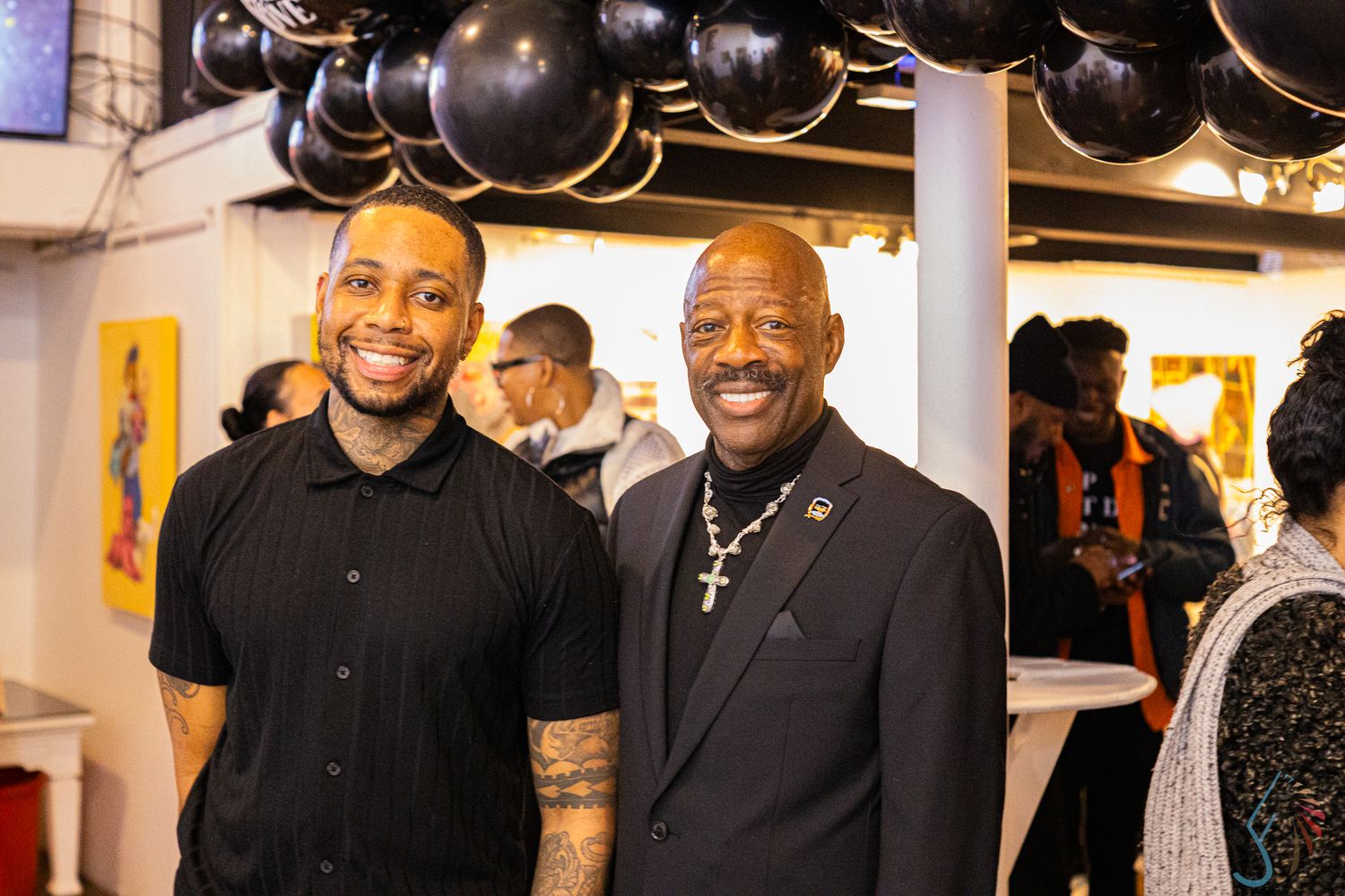 Two men smiling together at an art gallery event with black balloon decorations.